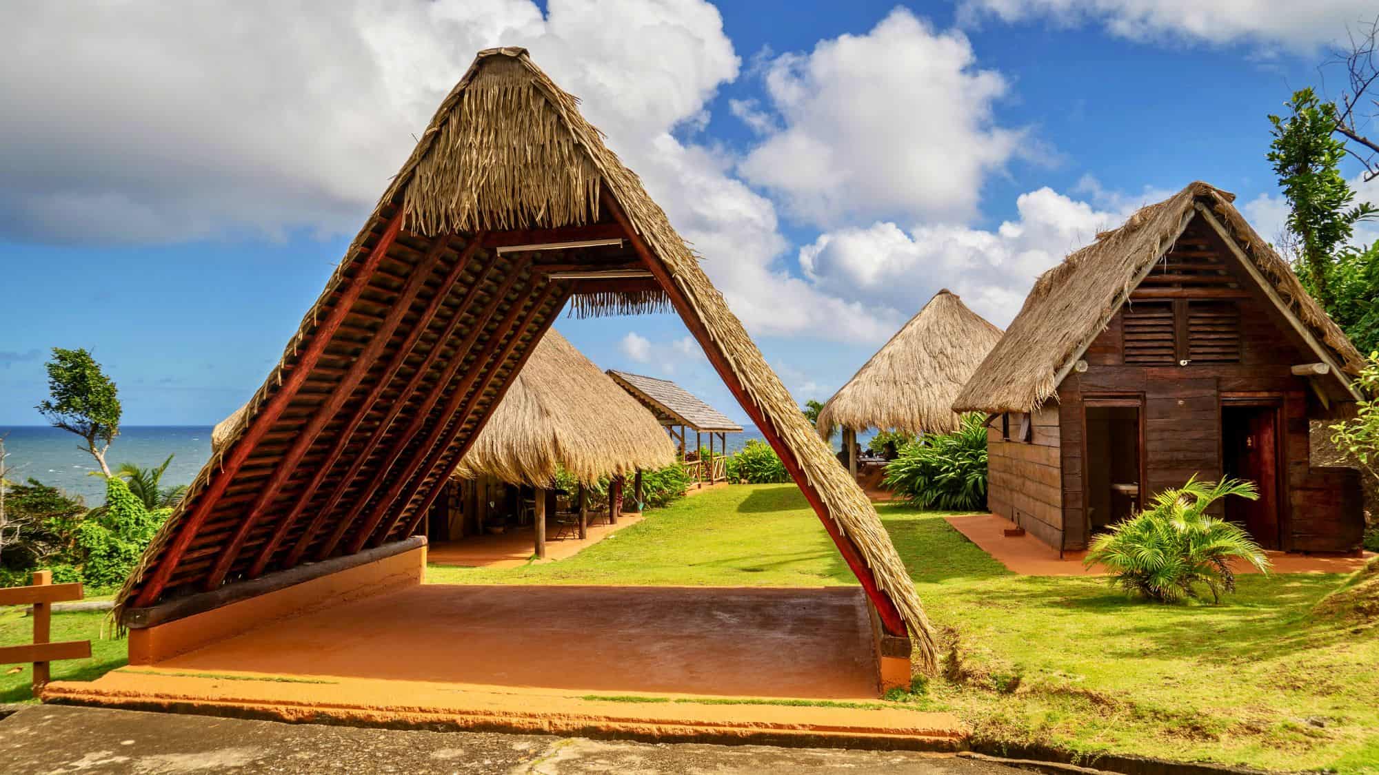 Traditional A-frame thatched huts in a seaside cultural village, surrounded by manicured green grass and tropical landscaping under a partly cloudy sky.