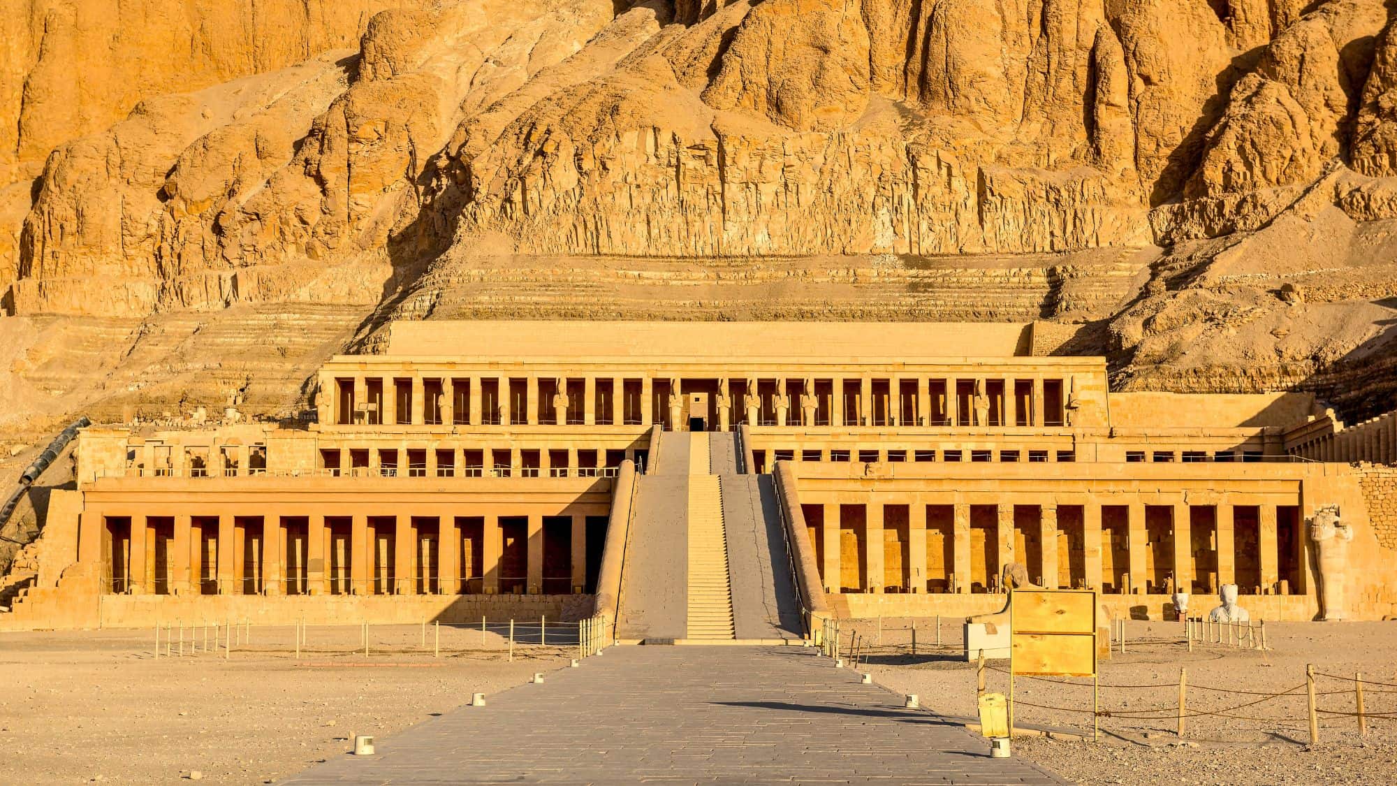 A symmetrical and grand structure carved into a desert cliffside in Luxor, Egypt, showing the temple of Hatshepsut with rows of stone columns and a central ramp leading upward.
