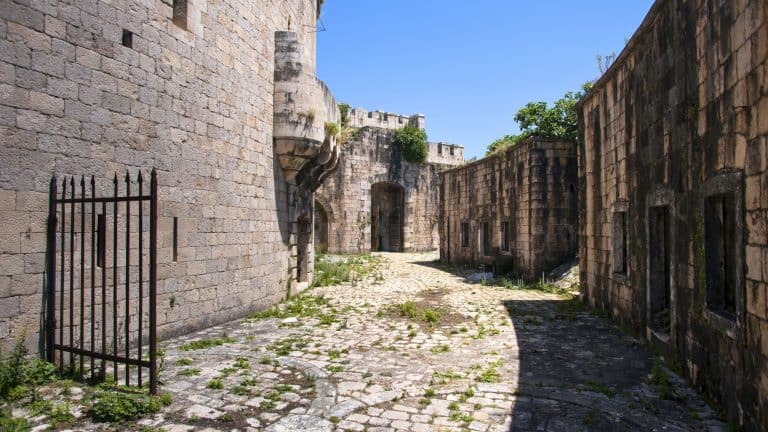 A sunlit cobblestone courtyard winds between high stone walls of the Mamula fortress, with empty window frames and a partially open black iron gate hinting at the structure’s abandoned state. Vegetation peeks through the cracks, adding a touch of nature to the historical ruin.