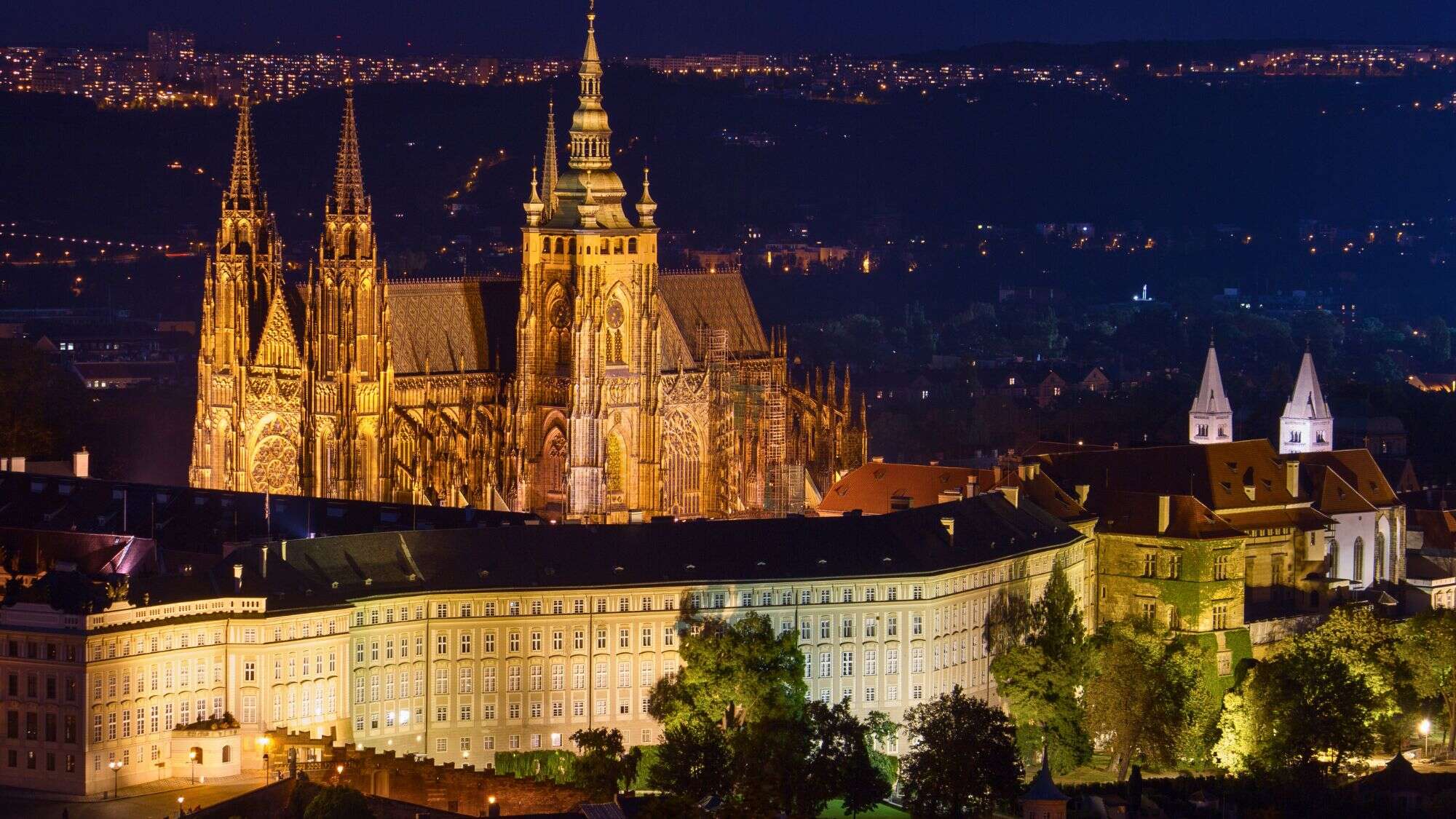 A majestic nighttime view of Prague Castle illuminated in golden tones, showcasing its Gothic spires and detailed architecture, with the city lights twinkling in the background.