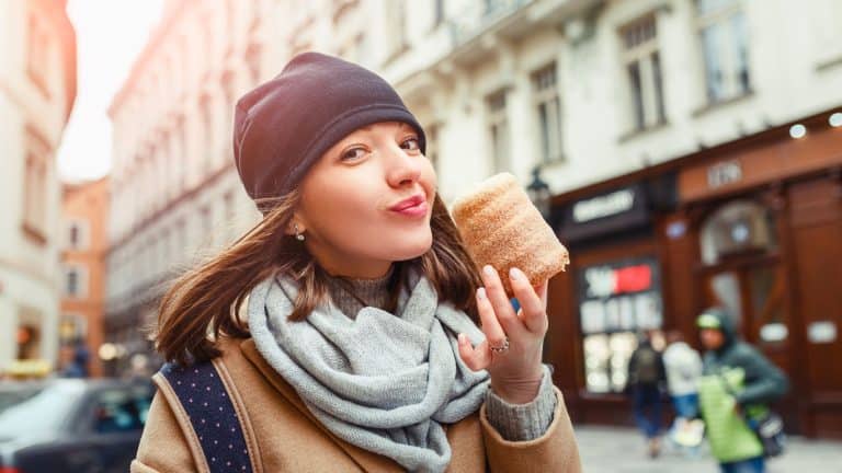 A woman in a scarf and beanie smiles while enjoying a freshly baked trdelník pastry on a cobblestone street, with blurred historic buildings in the background.