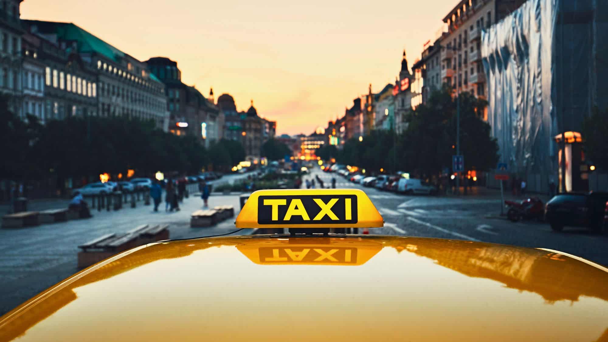 Close-up of a yellow taxi sign on a car roof at sunset, set against a bustling street lined with historic buildings and glowing city lights in Prague.