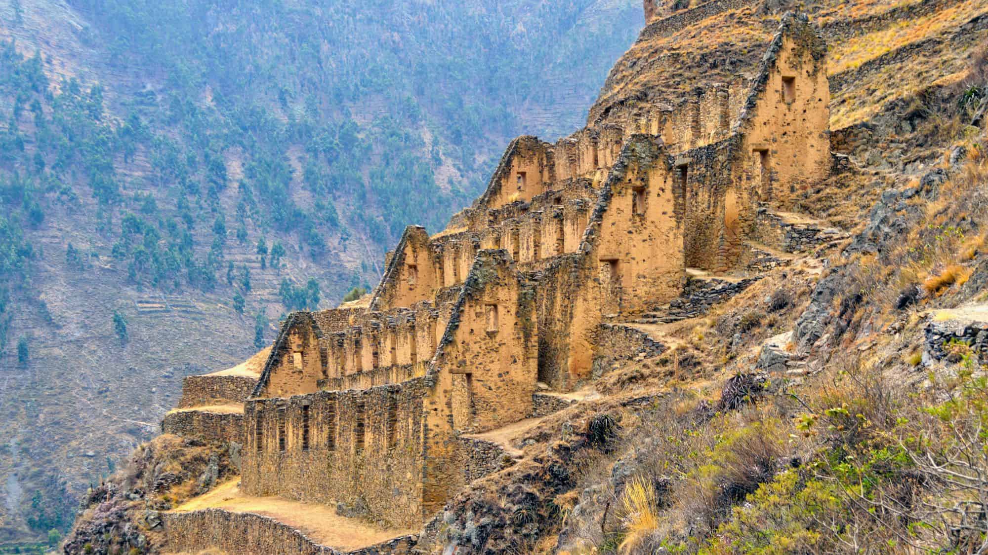 Sloped Incan stone terraces in Peru's Sacred Valley with crumbling, golden-brown walls set against a misty mountain backdrop covered in sparse vegetation and trees.