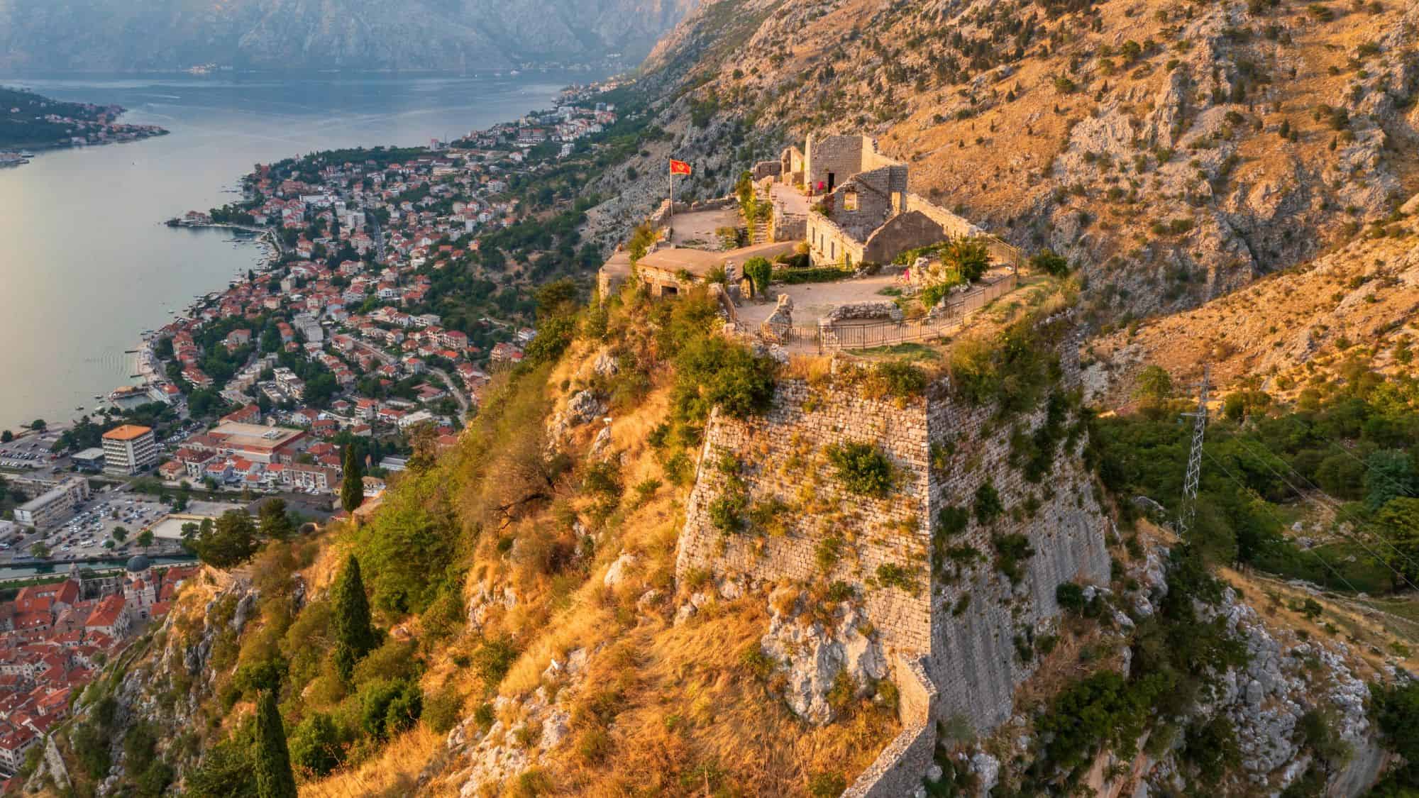 Dramatic aerial view of San Giovanni Fortress above Kotor, Montenegro, with steep fortress walls hugging the hillside and a panoramic look at the Bay of Kotor and town below.