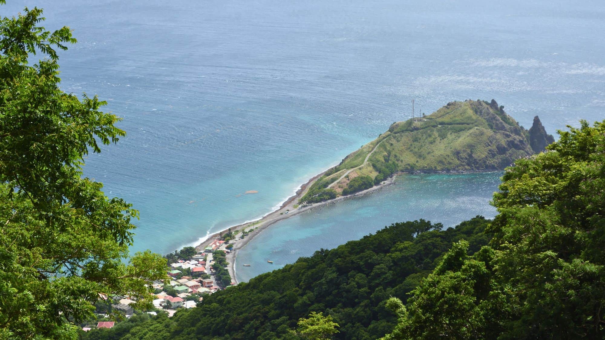 Scenic aerial view of a narrow strip of land extending into the ocean, with a small coastal village below and the surrounding turquoise sea framed by thick trees.