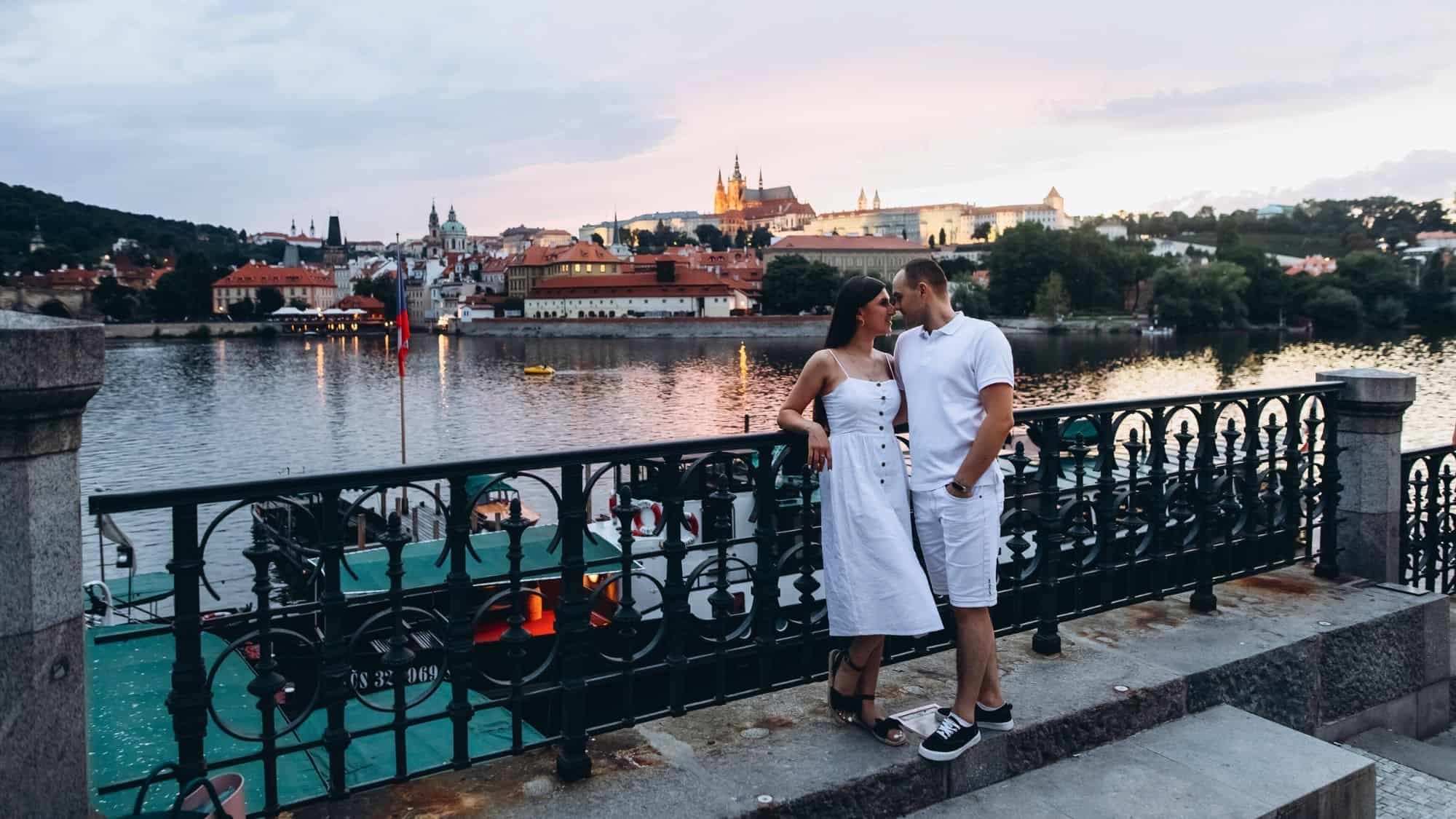 A couple in white stands close together on a riverside promenade in Prague at dusk, with Prague Castle and historic buildings lit in the background.