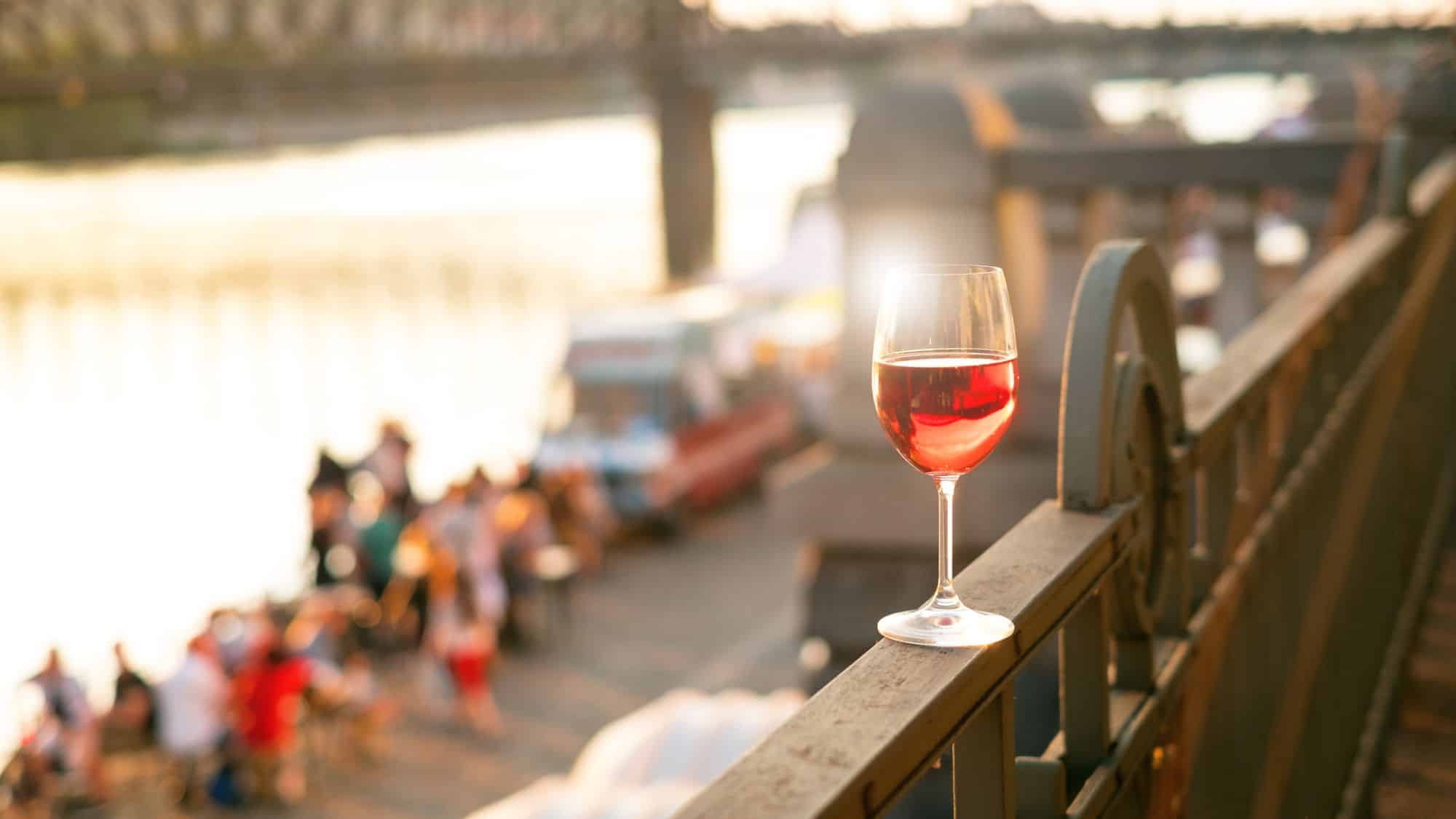 A single glass of rosé wine rests on a railing in soft golden light, with blurred people, food trucks, and a riverbank scene in the background.
