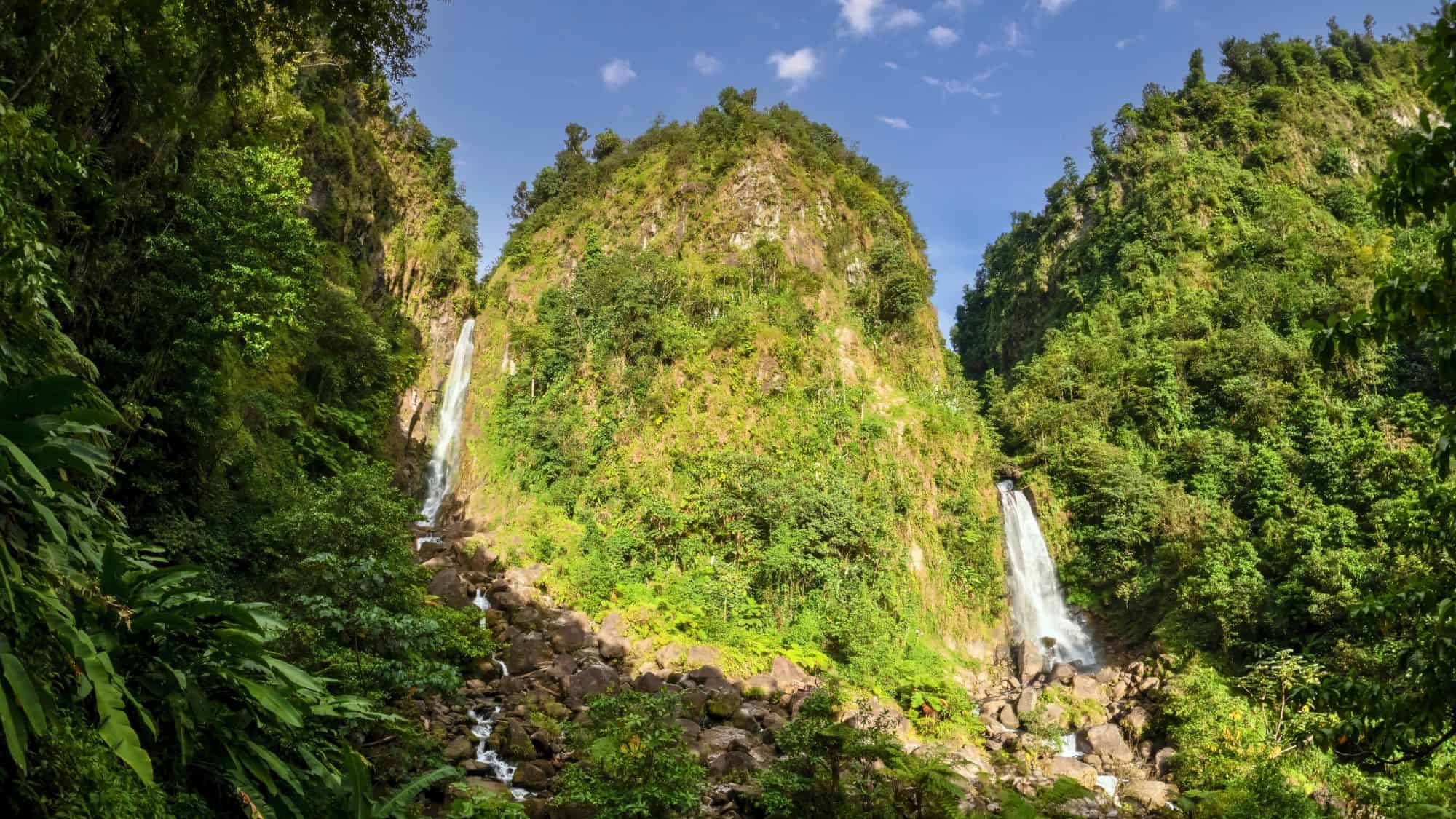 Twin waterfalls drop from lush green cliffs into a rocky basin, surrounded by thick rainforest in this iconic natural site within Dominica’s interior.