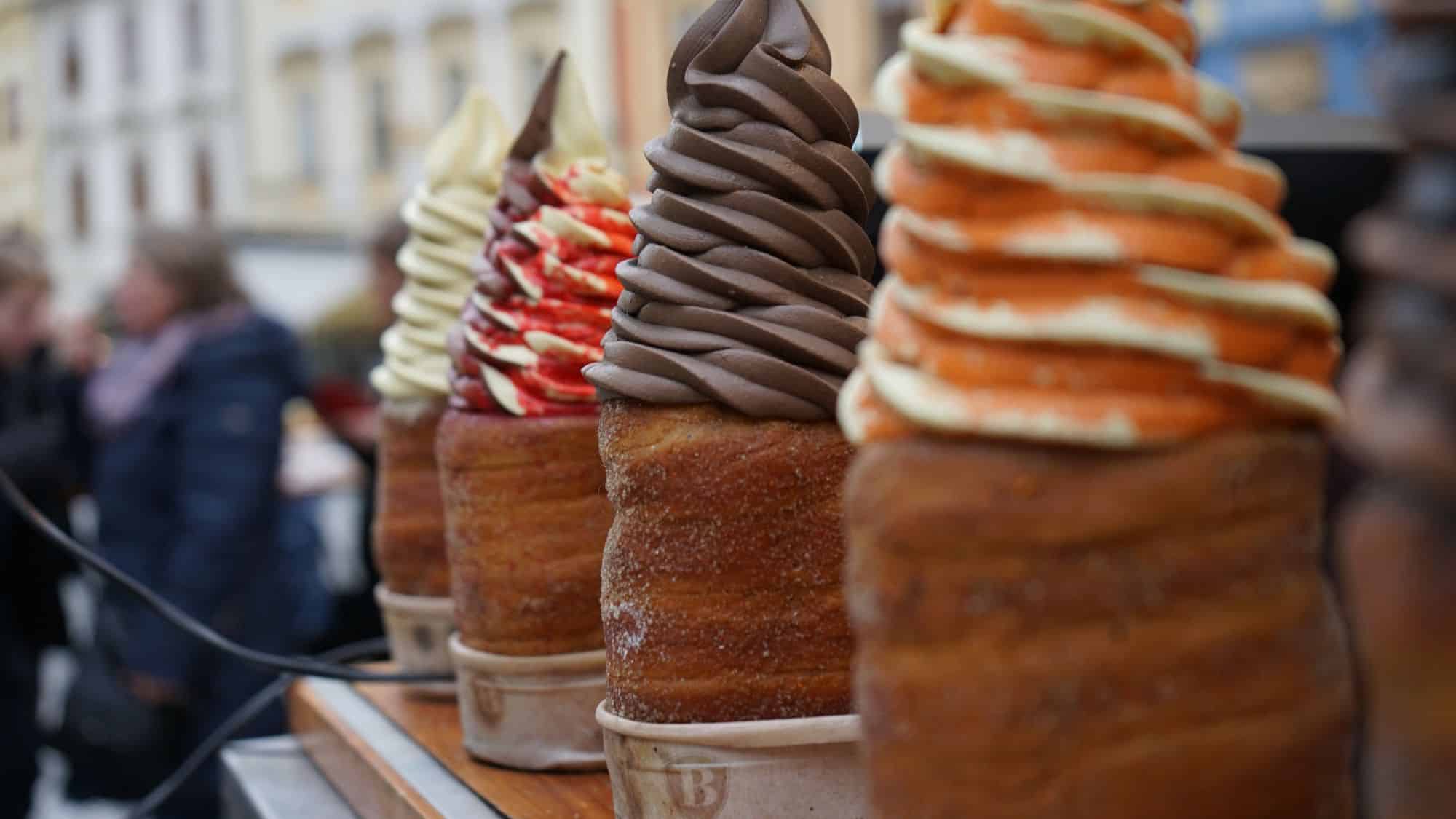 Close-up of colorful ice cream swirls served in traditional chimney cakes (Trdelník), lined up at a street food stand in Prague.