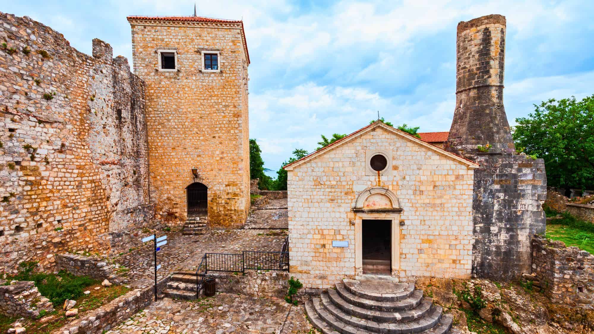 Stone fortress ruins in Ulcinj, Montenegro, featuring a small chapel with rounded steps and a square tower, surrounded by weathered medieval walls.