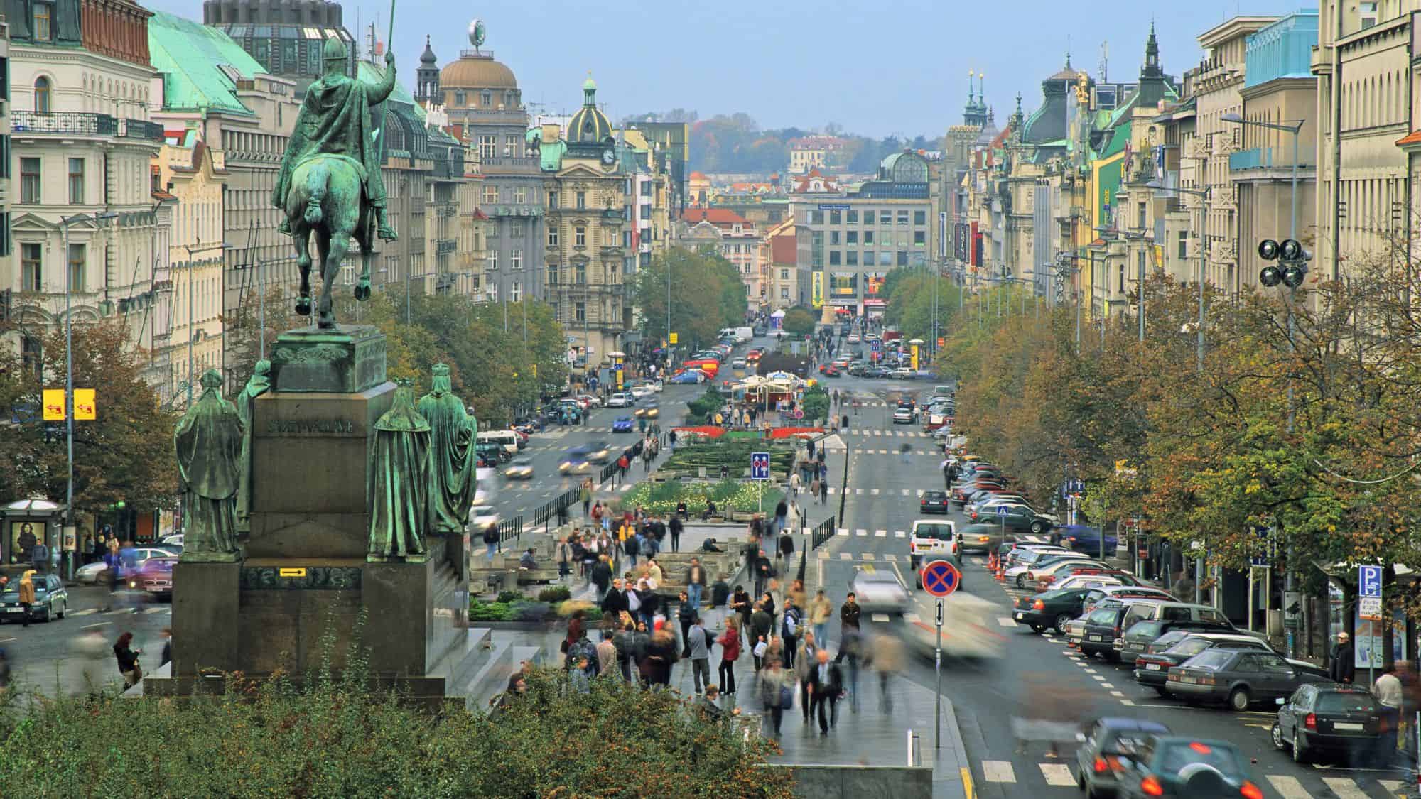 A wide view of Wenceslas Square from the base of the Saint Wenceslas statue, showing the long boulevard filled with pedestrians, traffic, and classic Prague architecture.