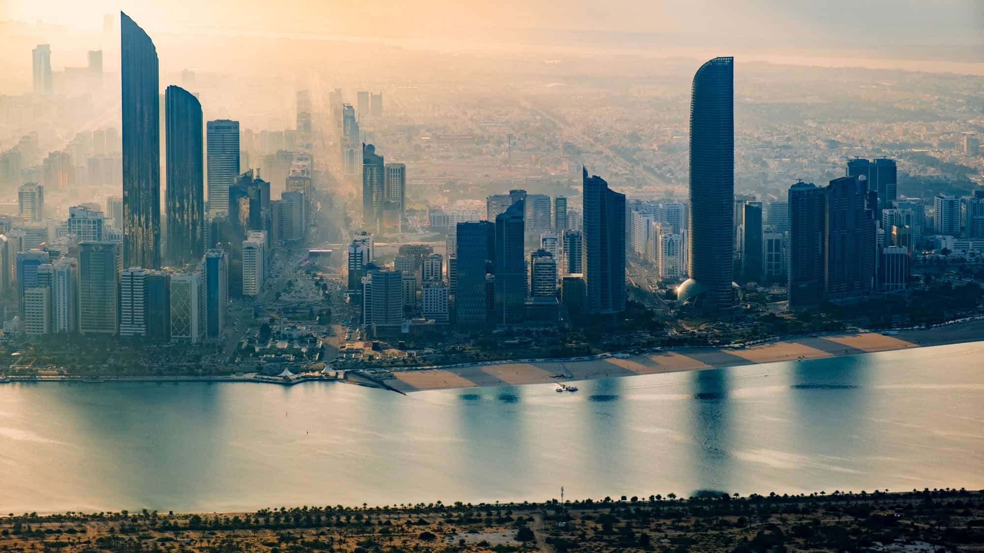Dramatic aerial view of Abu Dhabi's skyline at dusk, with soft golden light casting long shadows from sleek skyscrapers, while the calm waters of the Persian Gulf reflect the hazy silhouette of the city.