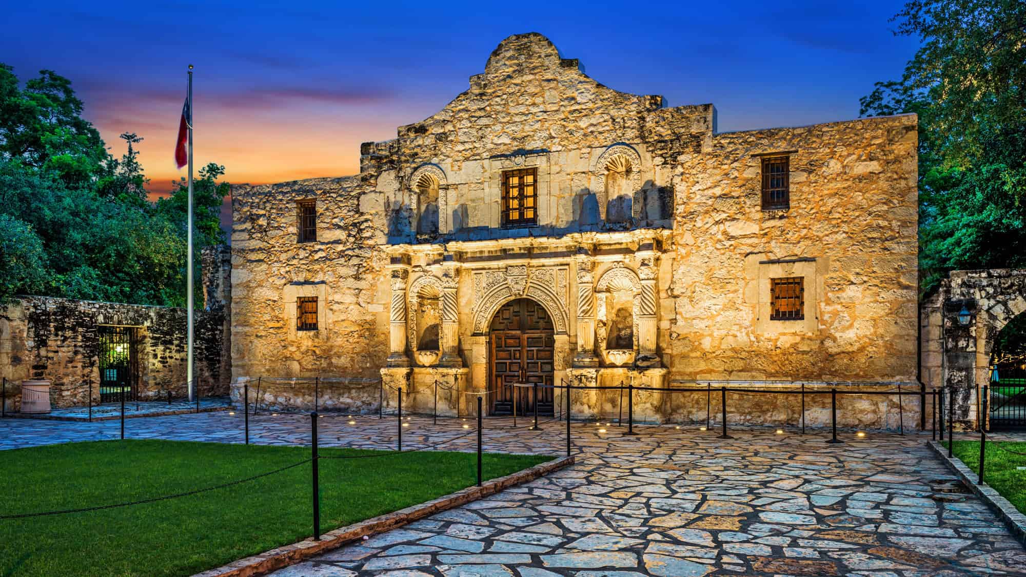 Iconic limestone mission building with Spanish colonial architecture, illuminated at dusk with dramatic blue and orange skies.