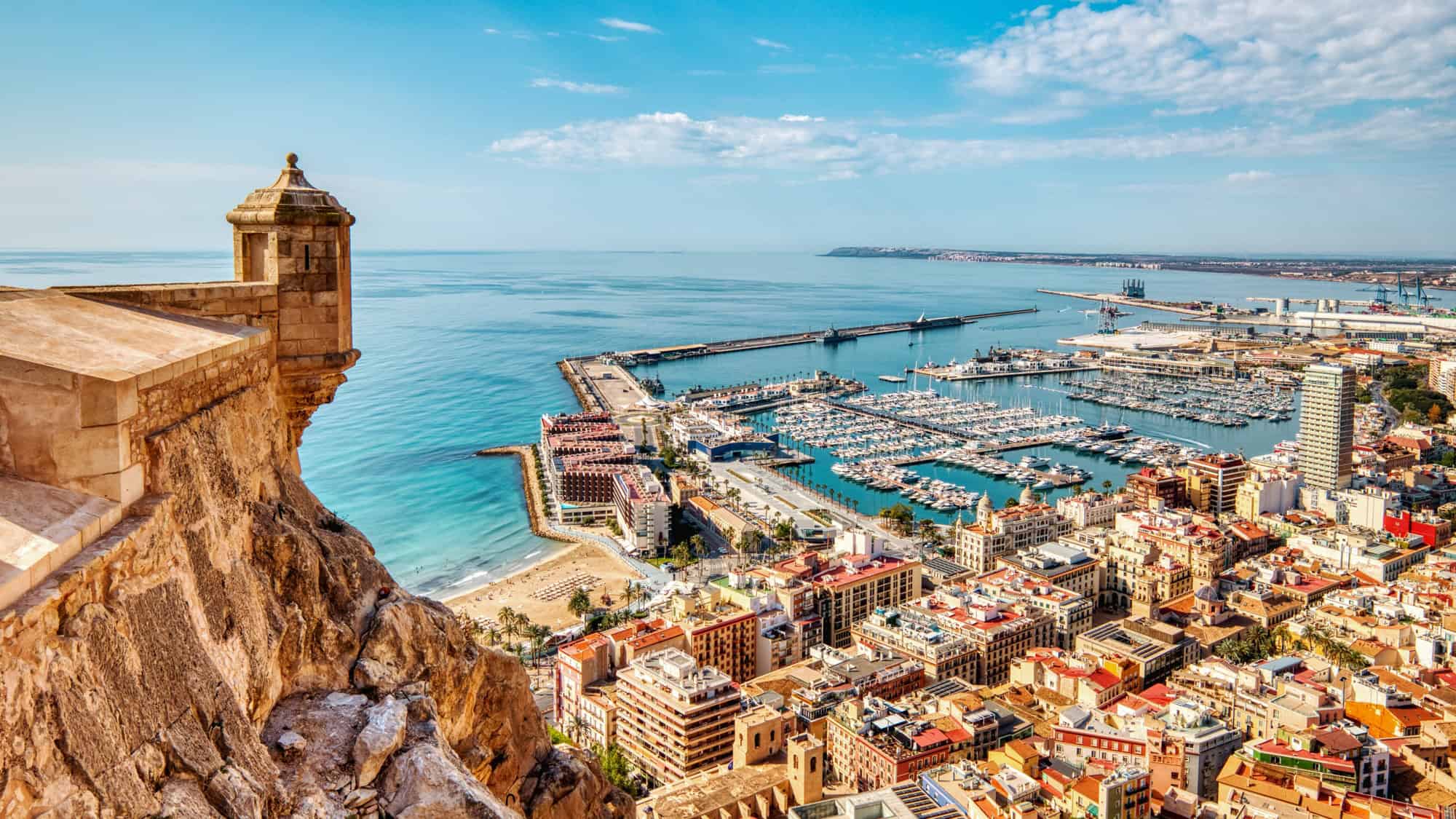 Sun-drenched aerial view of Alicante, Spain, showing a historic fort wall perched on a rocky cliff overlooking a bustling marina full of boats and the deep turquoise waters of the Mediterranean.