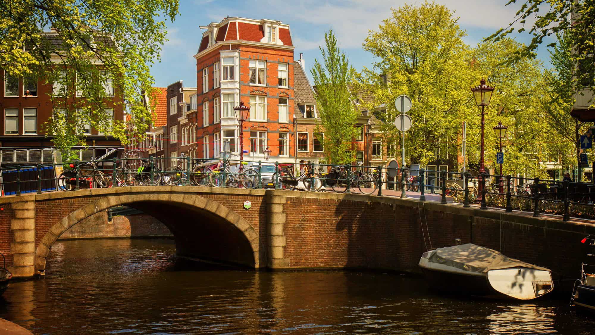 A charming canal bridge in Amsterdam, lined with parked bicycles and traditional Dutch buildings, with a small boat moored beneath and spring greenery all around.