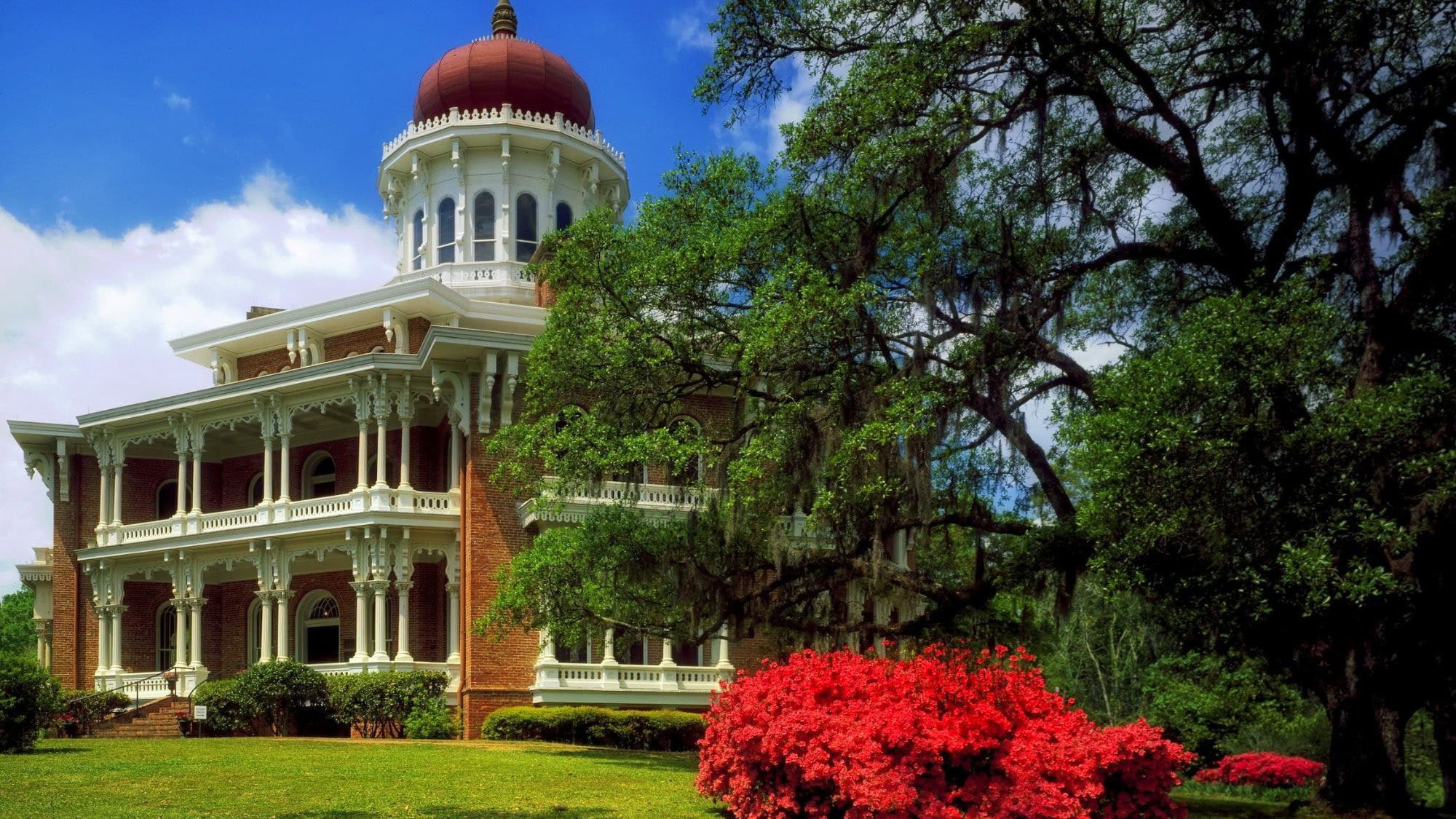 A grand Southern mansion with red brick walls, intricate white columns, and a large white dome with a red cap, surrounded by flowering azaleas and draped oak trees in full bloom under a bright blue sky.