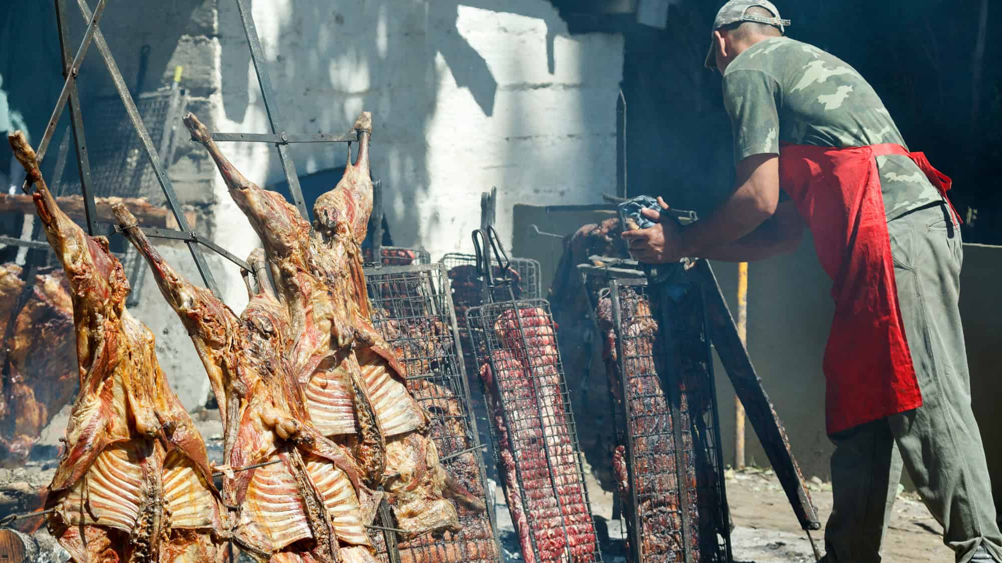 A man in a red apron tends to large racks of meat cooking over open flames in a traditional outdoor asado.