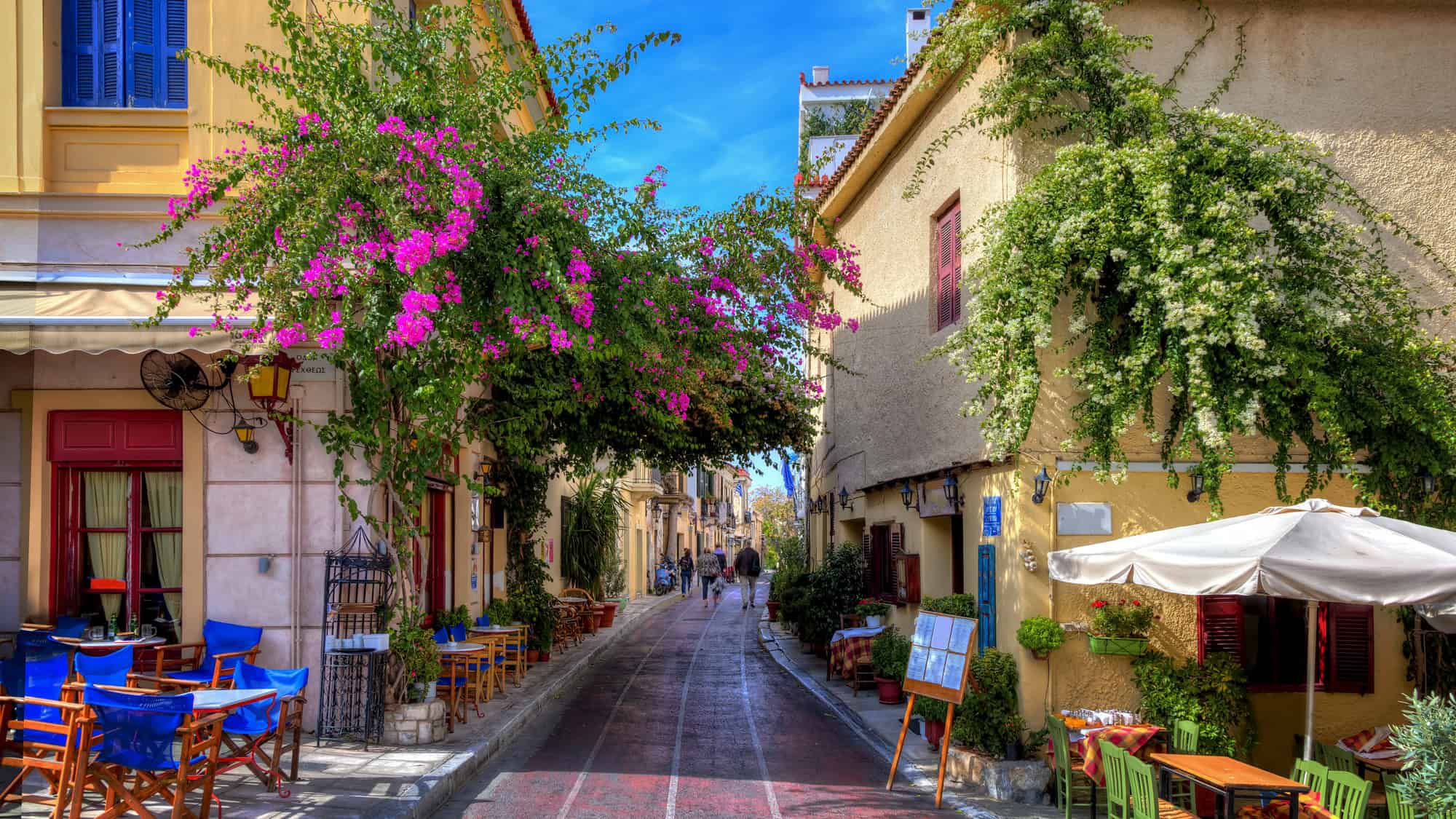 A charming pedestrian lane in Athens’ Plaka district is lined with colorful buildings, flowering bougainvillea, and inviting sidewalk cafés with bright blue chairs and tables under a vivid blue sky.