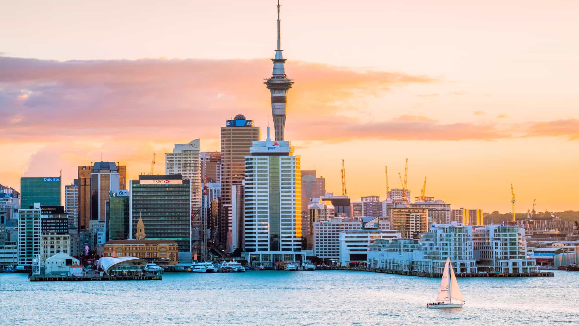 Evening view of Auckland, New Zealand’s waterfront with the Sky Tower at center, surrounded by high-rise buildings and cranes, as a white sailboat glides across the water beneath a colorful sunset sky.