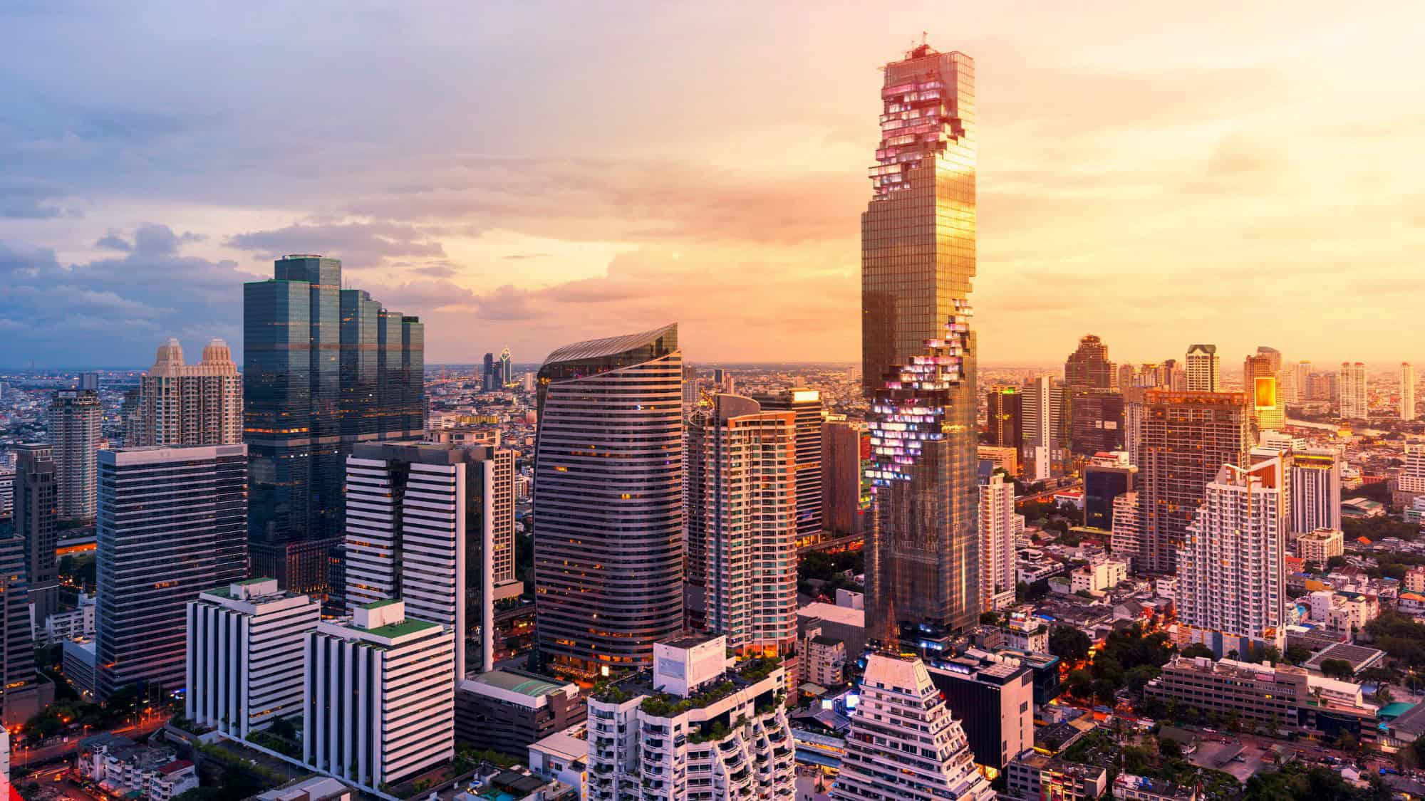 A golden sunset bathes Bangkok’s cityscape, featuring a mix of futuristic skyscrapers, including the pixelated MahaNakhon tower, rising above the dense urban landscape.