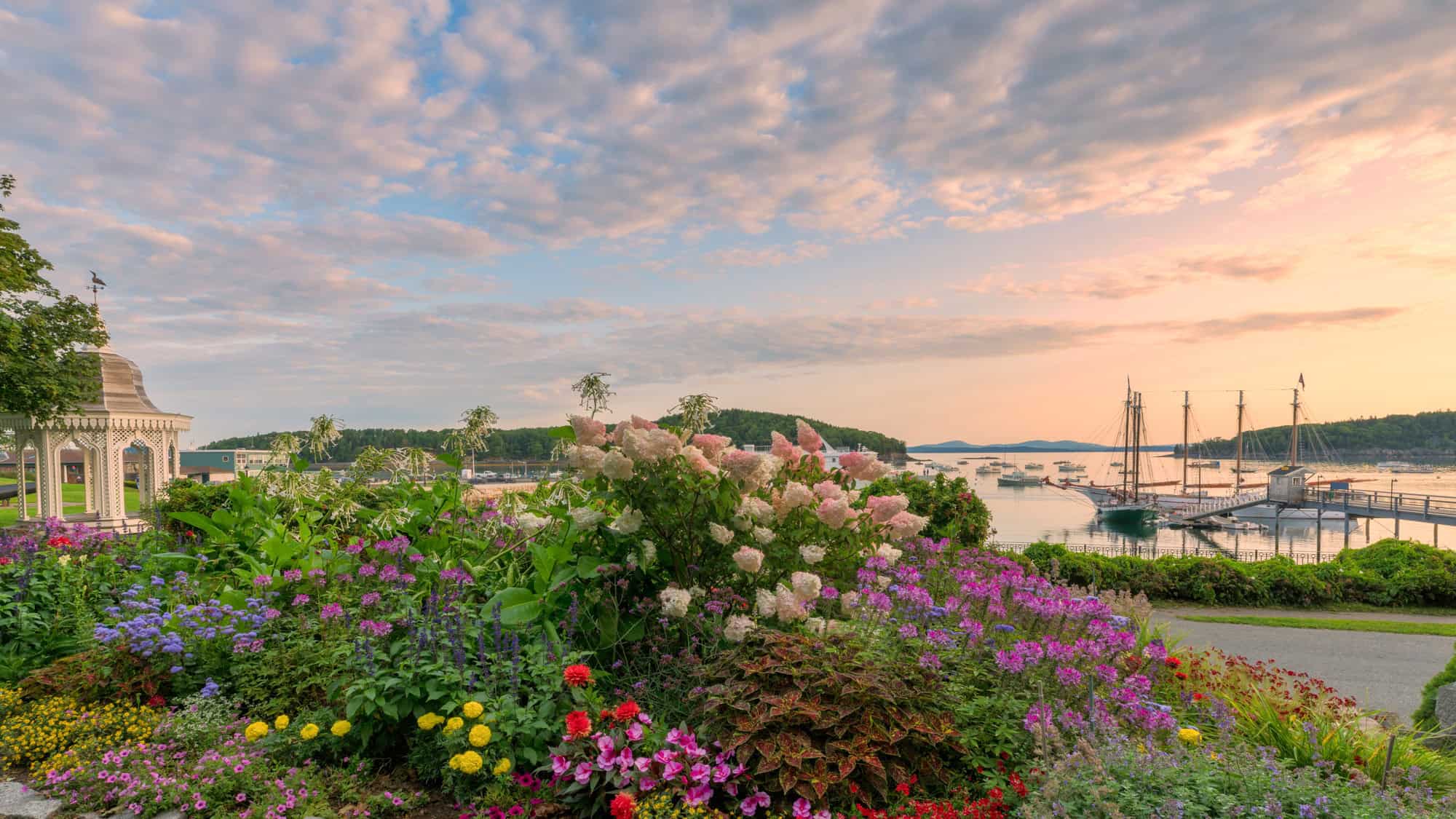 A vibrant flower garden blooms in the foreground with a charming white gazebo to the left, while tall-masted schooners rest at the docks under a pastel-hued sunset sky in Bar Harbor, Maine.