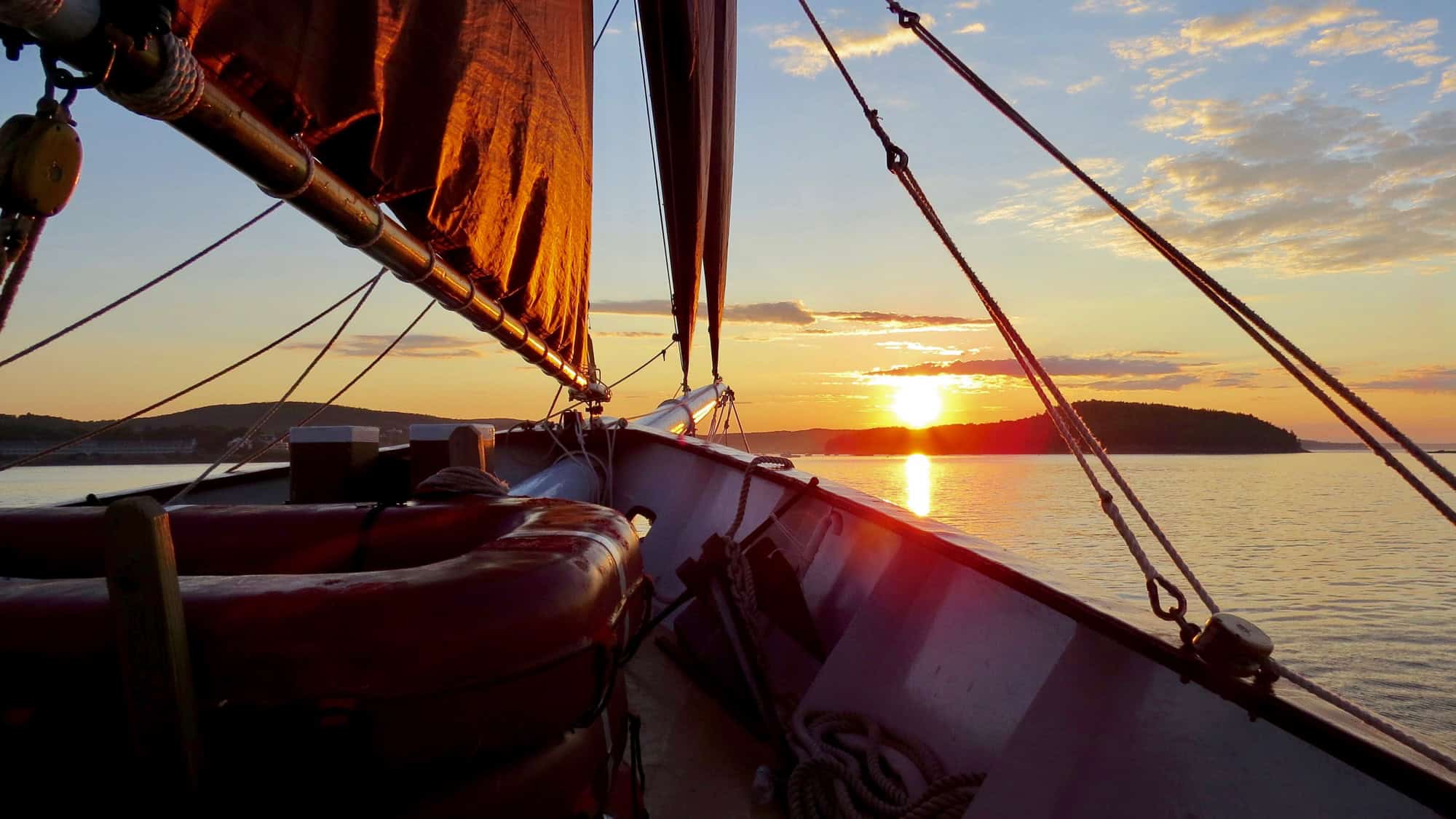 View from a sailboat at sunset, with the mast, ropes, and deck equipment silhouetted against the setting sun over calm coastal waters and low islands in the distance.