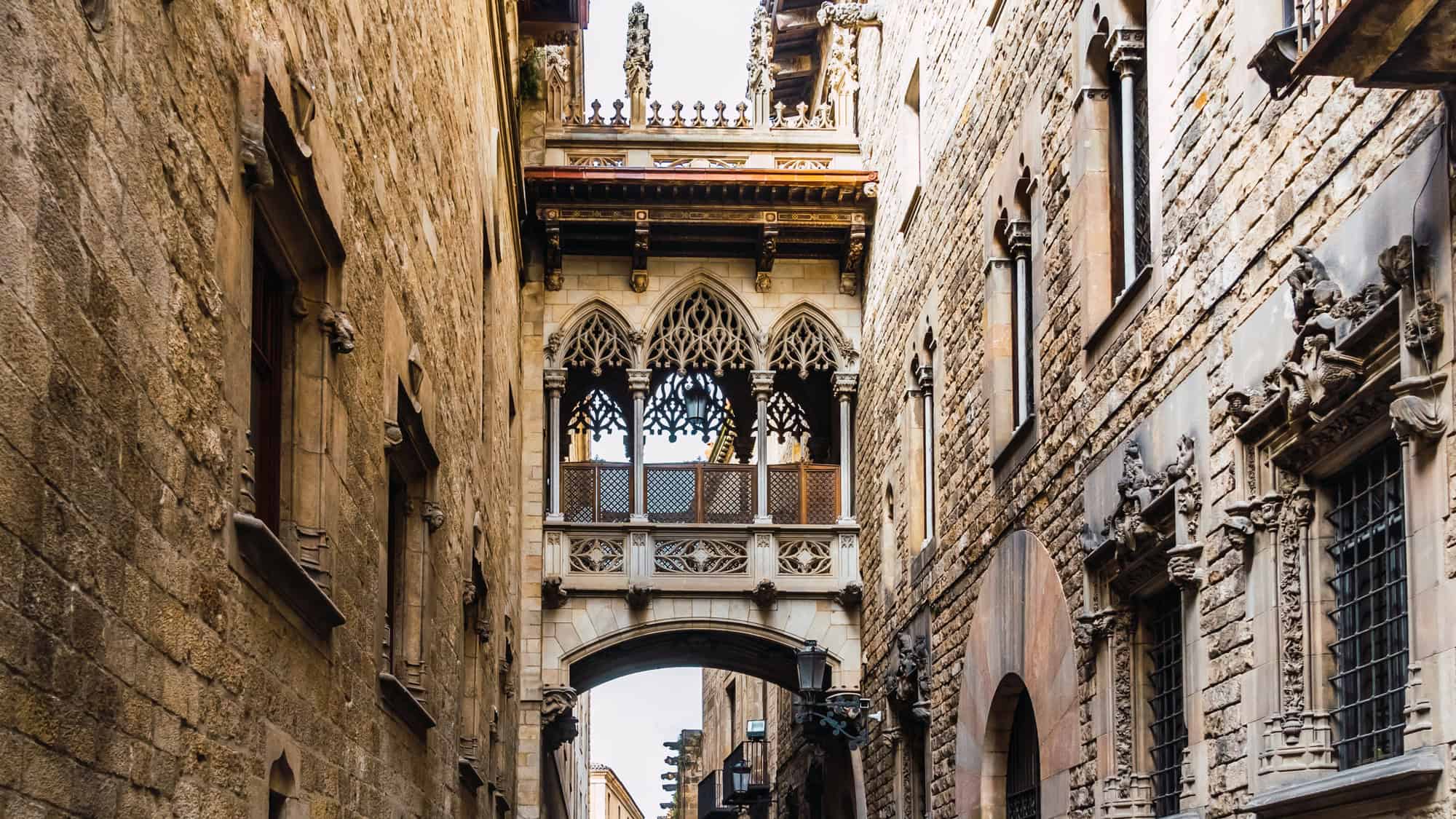 A stone footbridge with ornate gothic details connects two historic buildings in Barcelona’s Gothic Quarter. Pointed arches and intricate latticework give the scene a medieval, architectural elegance.