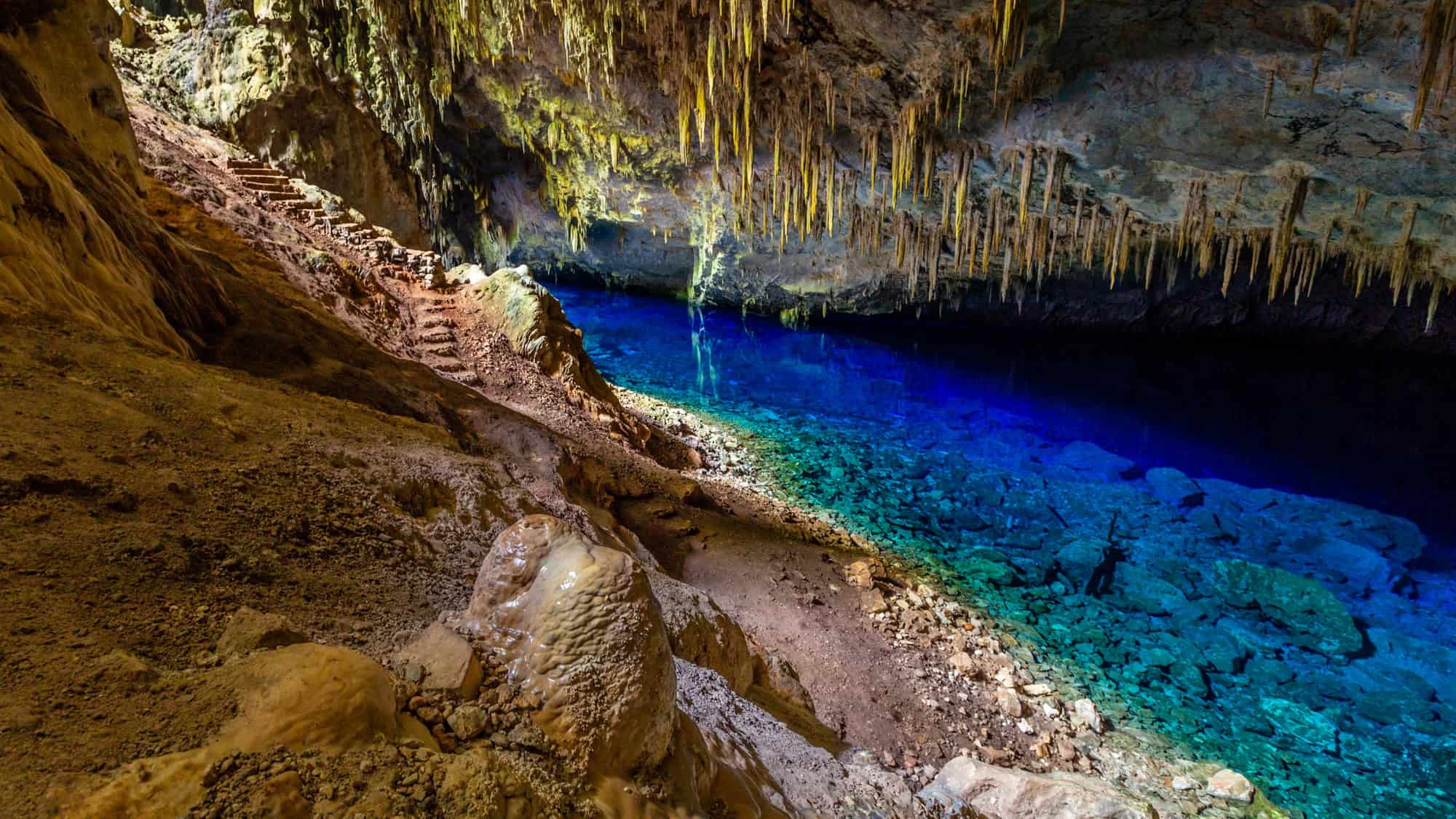 An underground cave in Brazil’s Mato Grosso do Sul featuring a glowing blue lake surrounded by rock formations, stalactites, and a natural stone staircase.