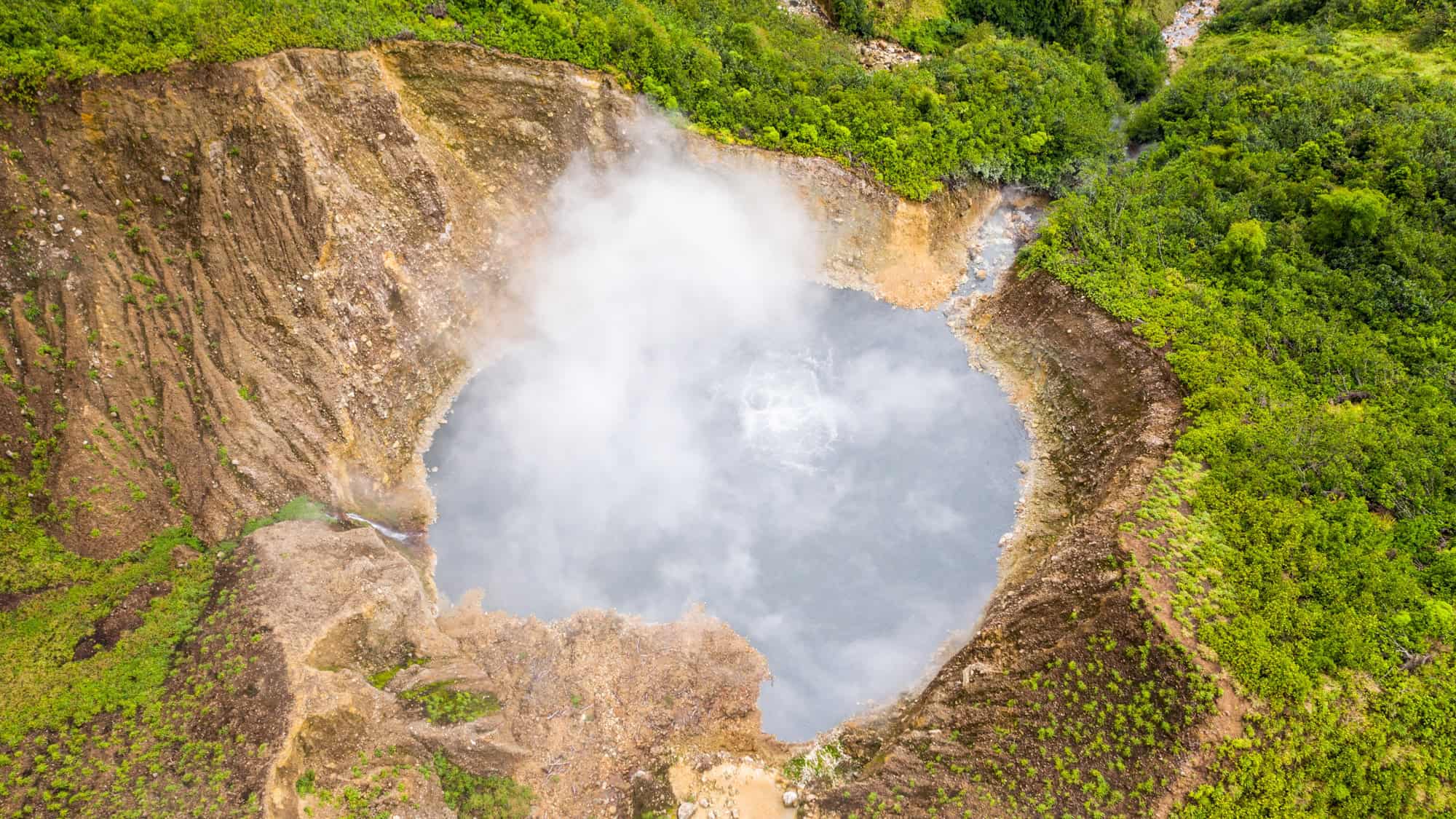 A wide crater filled with pale gray-blue boiling water, surrounded by steep rocky cliffs and dense mist, creating a mysterious and dramatic volcanic landscape.