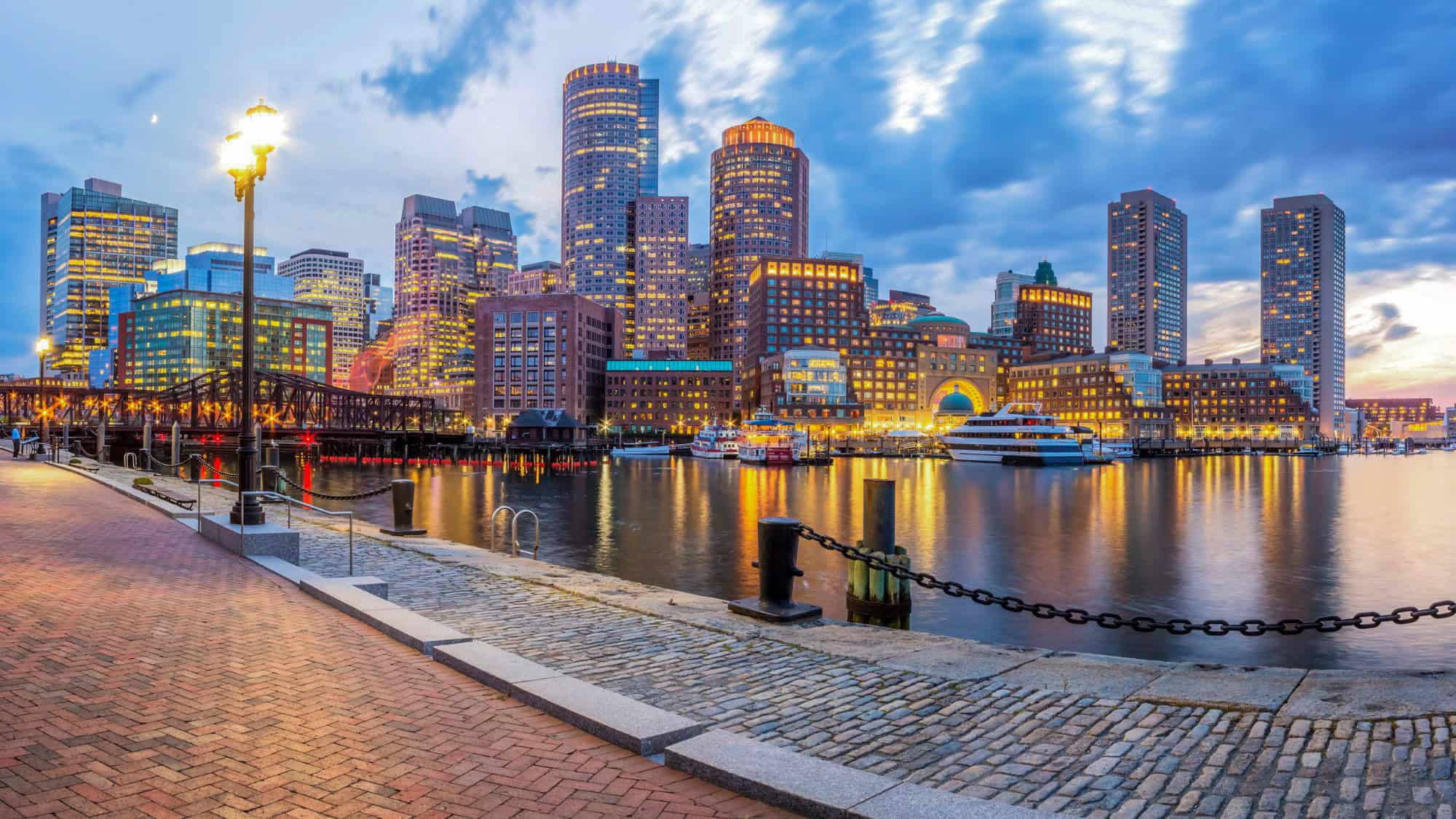 A glowing city skyline reflects in the calm waters of Boston Harbor as twilight sets in, with boats docked and streetlamps lighting the cobblestone promenade.