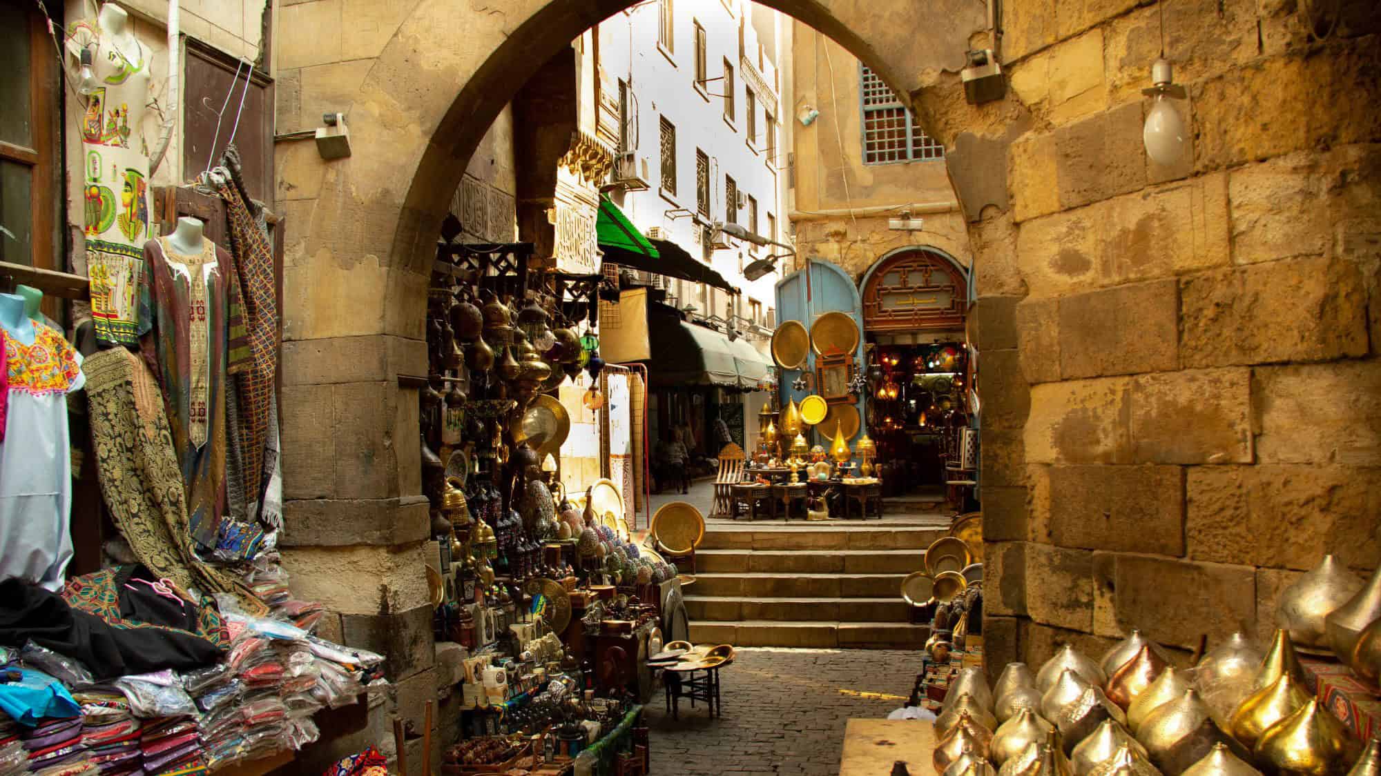 A historic bazaar in Cairo is packed with brass lamps, textiles, and colorful garments under an archway. Sunlight filters in, highlighting the intricate patterns and old stonework of the market walls.