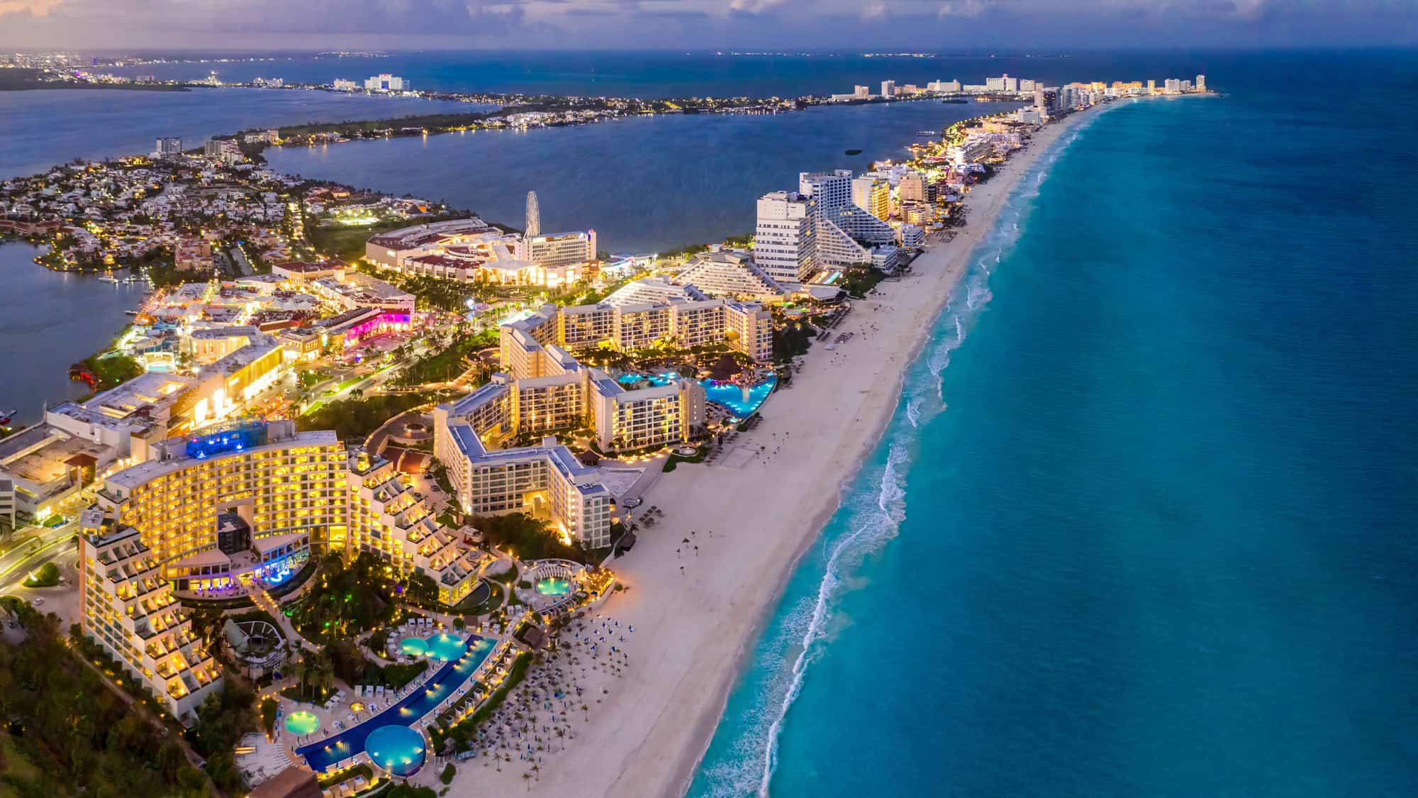 A panoramic aerial view of Cancun’s Hotel Zone at twilight, with luxury resorts glowing along the white sand beach and the turquoise Caribbean Sea stretching to the horizon.