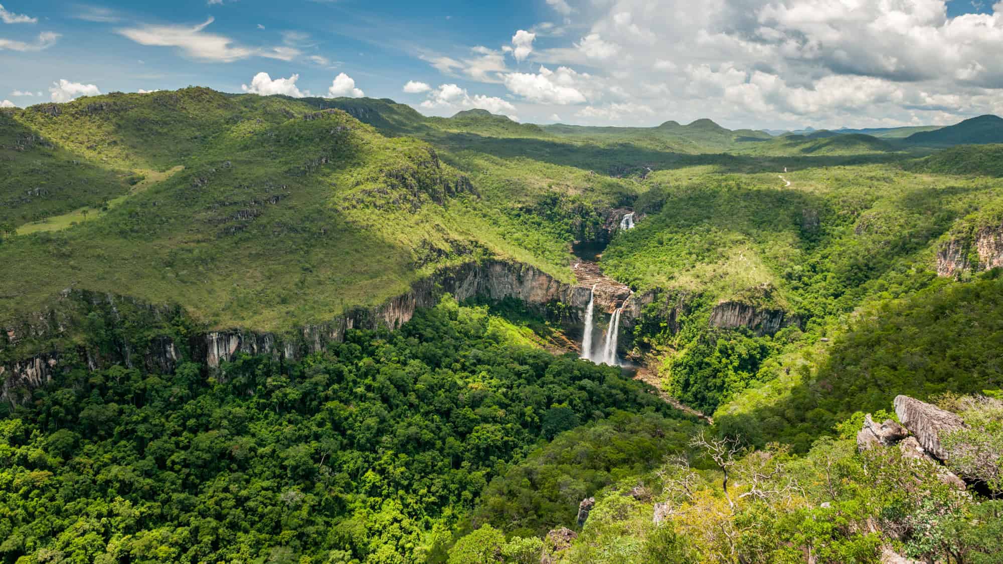 A dramatic landscape of lush green mountains and plateaus with twin waterfalls plunging into a canyon in Chapada dos Veadeiros National Park, Brazil, under a partly cloudy sky.