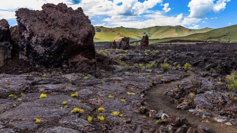 A rugged volcanic landscape with sharp lava rock formations, sparse vegetation, and a winding dirt path under a partly cloudy sky and green hills in the distance.