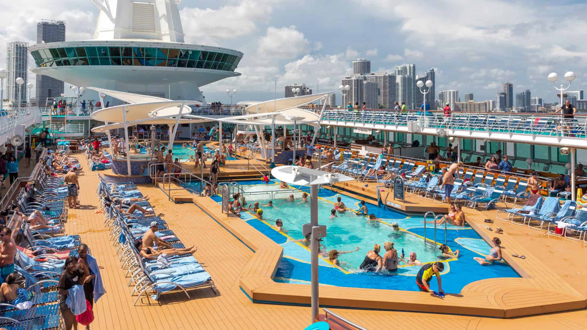 A bustling cruise ship deck scene shows guests relaxing and swimming near a pool surrounded by lounge chairs and city skyline in the background, under partly cloudy skies.