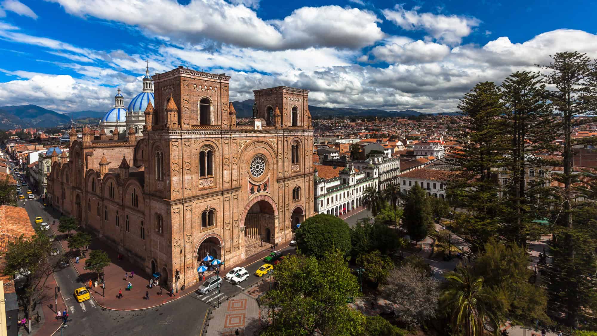 A majestic aerial shot of Cuenca, Ecuador, with the grand New Cathedral dominating the foreground—its terracotta towers and vibrant blue domes contrasting with the colonial-style buildings and surrounding mountains.