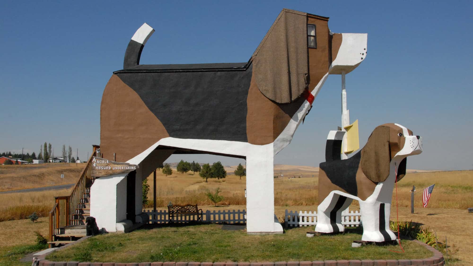 A whimsical roadside attraction shaped like a giant beagle serves as a guest inn in rural Idaho, complete with another smaller dog statue and a sign that reads, "A Noble Absurd Undertaking."