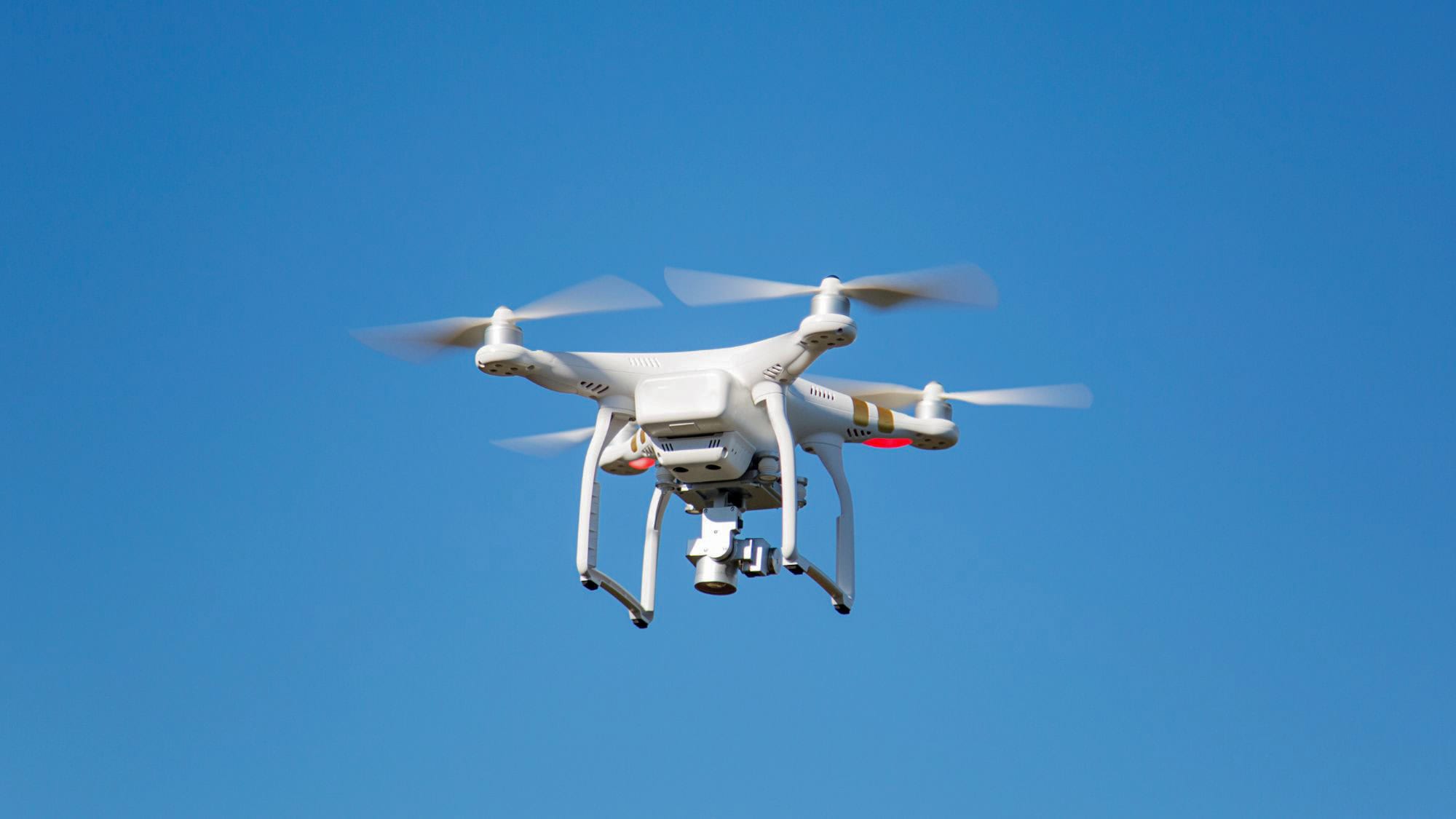 A white quadcopter drone hovers mid-air against a bright blue sky, its propellers blurred in motion, equipped with a camera mounted underneath.