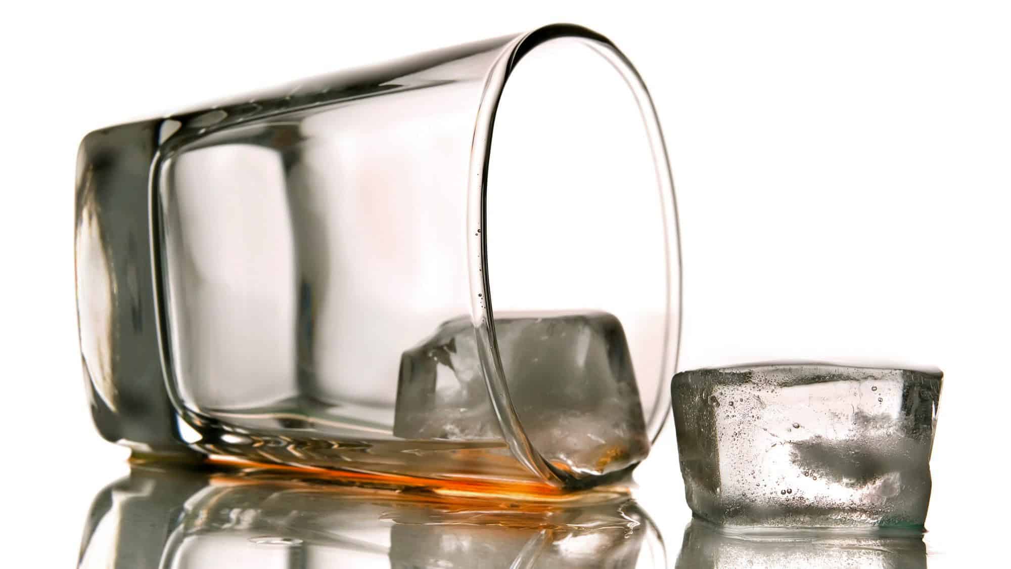 A tipped-over glass tumbler rests on a reflective surface with a couple of melting ice cubes, one still inside the glass with a splash of amber liquid remaining.
