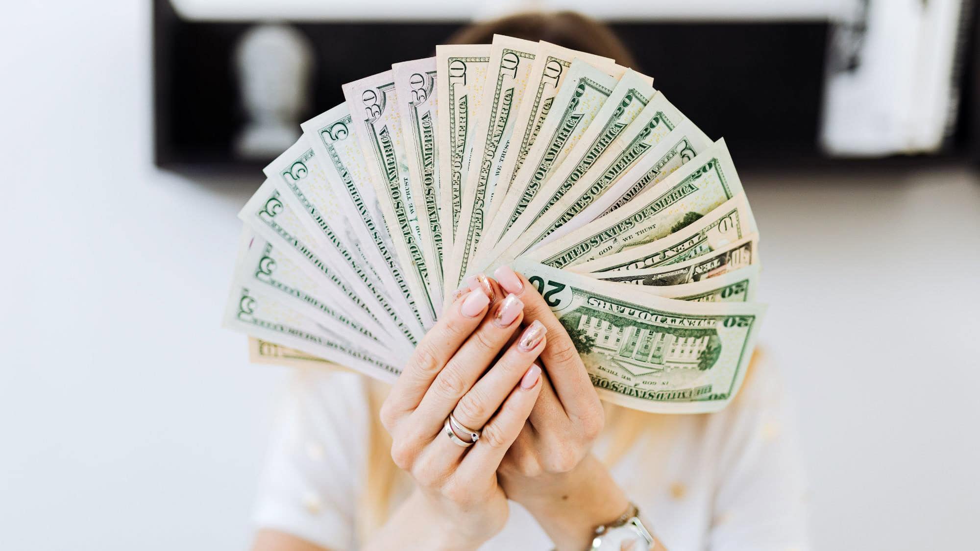 A woman’s manicured hands hold out a wide fan of U.S. dollar bills in various denominations, partially hiding her face, suggesting wealth or success.
