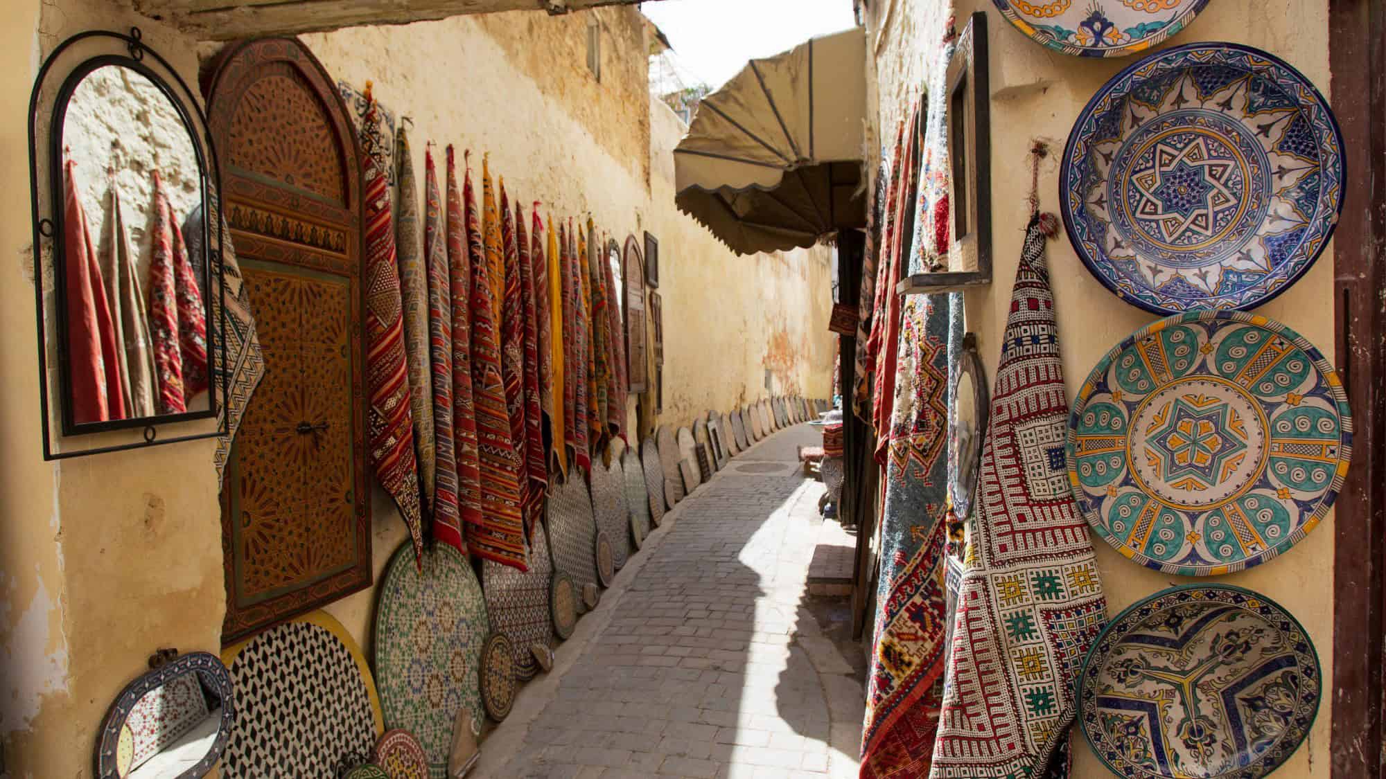 A narrow, sunlit alley in Fes, Morocco, lined with colorful rugs, ornate ceramics, and carved wooden decor, creating a vibrant and traditional marketplace scene.