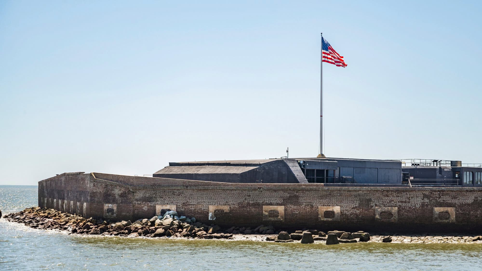 Low, historic stone fort surrounded by water with a large American flag flying atop, under a clear blue sky.