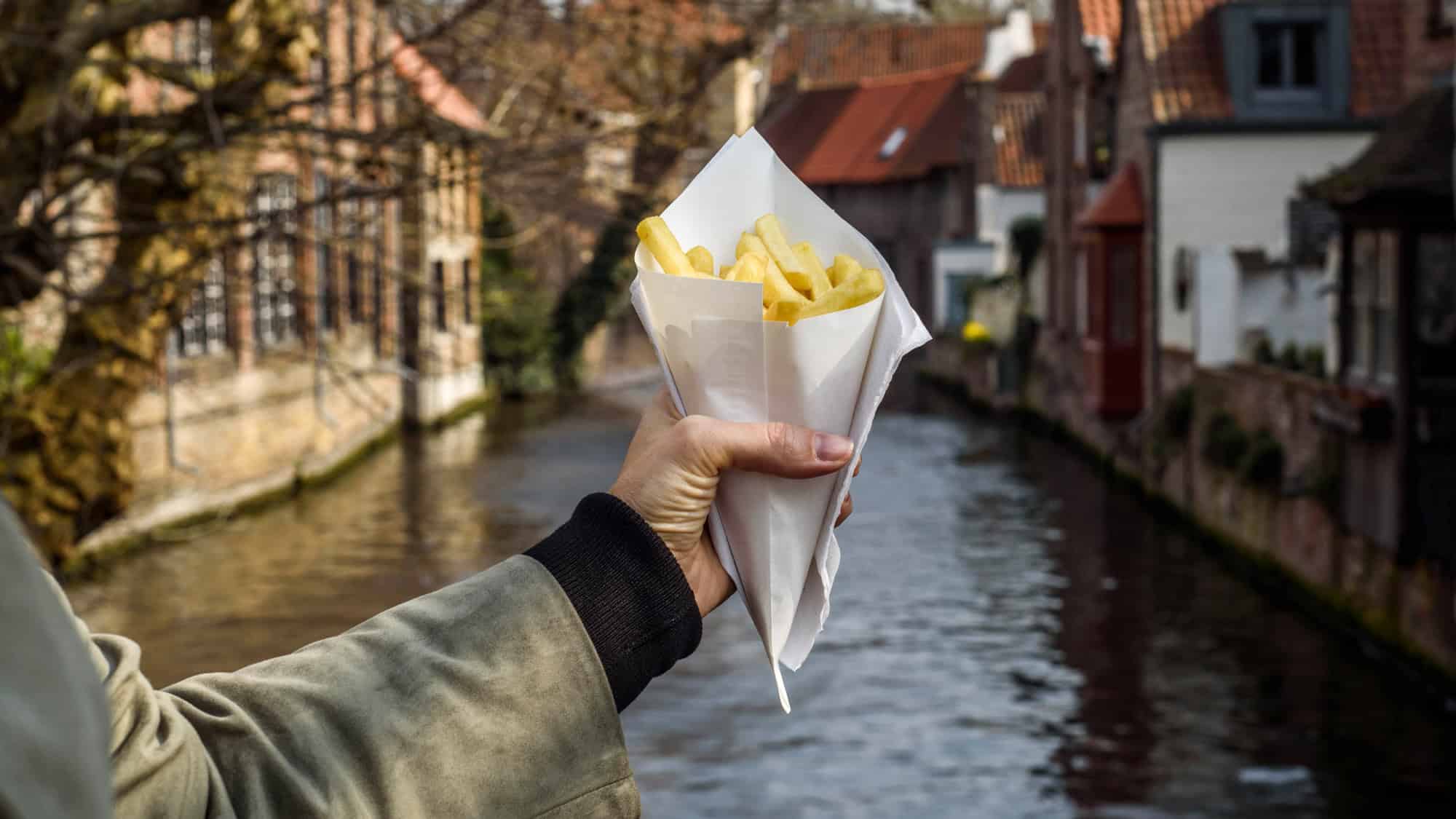 A hand holds a paper cone of golden fries against the backdrop of a scenic European canal and old brick houses.
