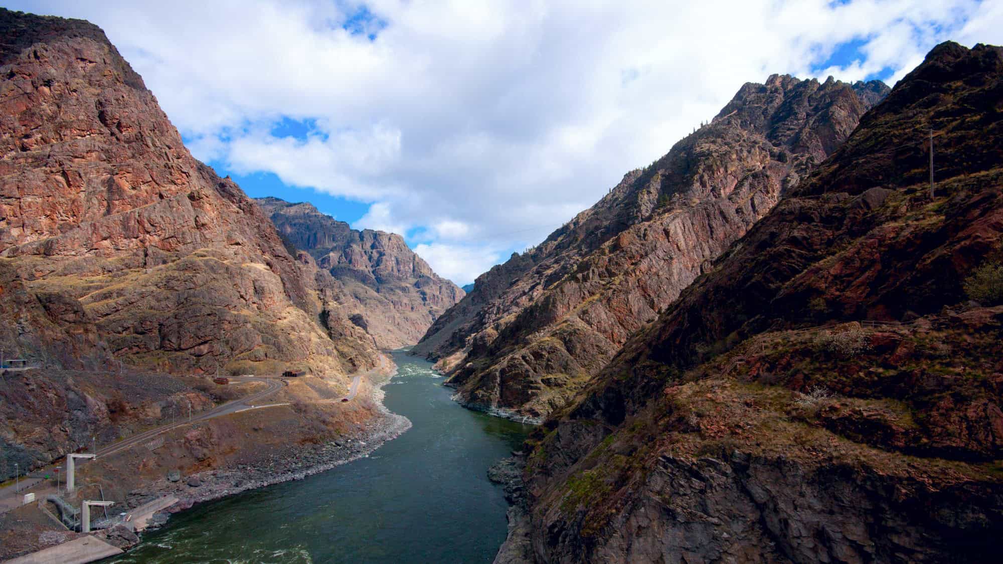 A deep, rugged gorge with steep, red-brown rock walls flanks the winding Snake River, showcasing the dramatic and remote beauty of Hells Canyon on the Idaho-Oregon border.