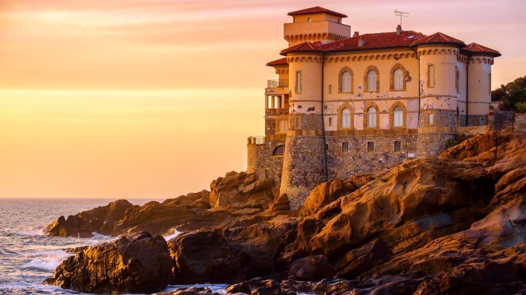 A grand, weathered villa sits dramatically on rocky cliffs above the sea at sunset, its stone base and terracotta roof glowing warmly in the fading light.