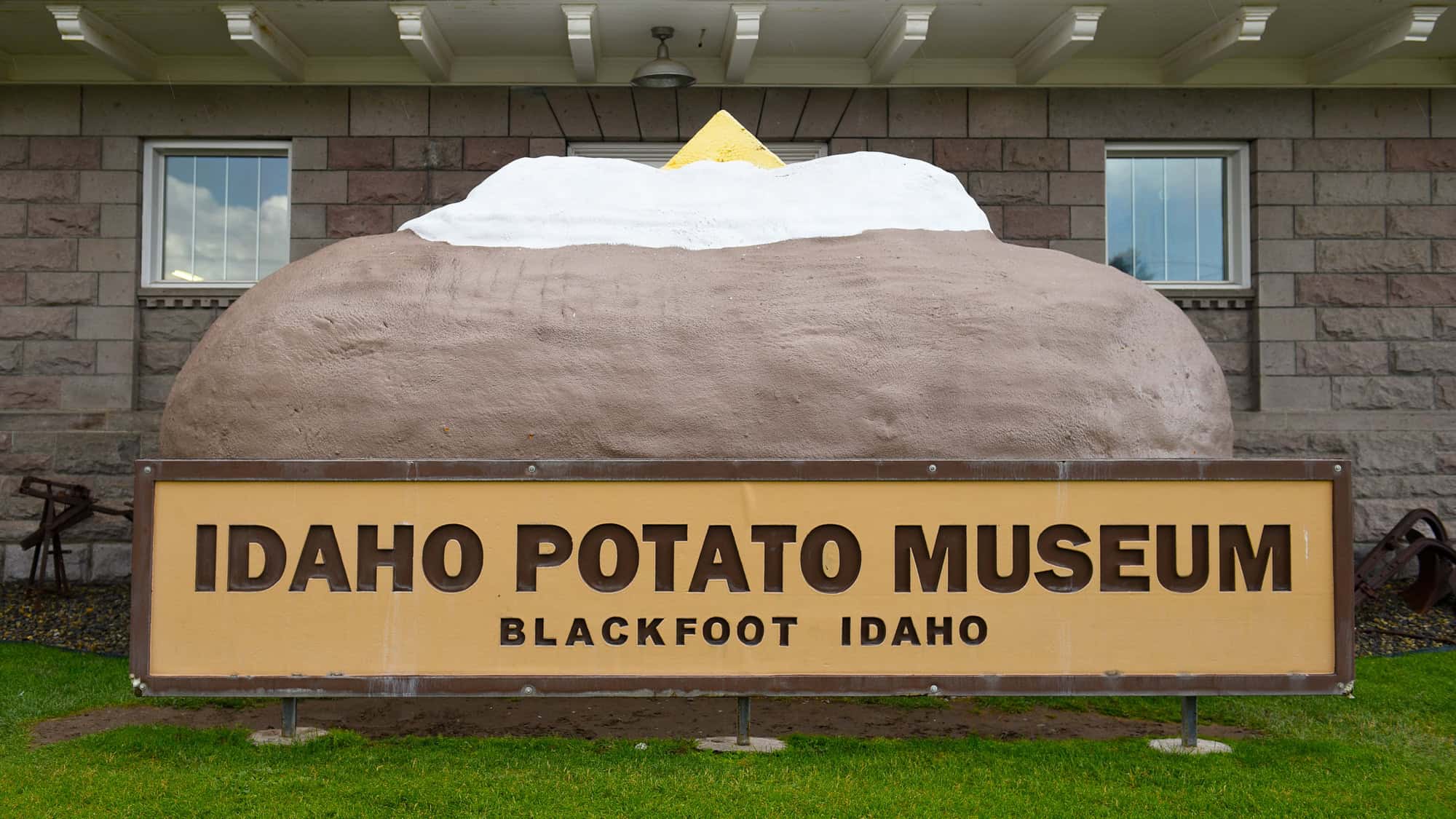 A large sculpture of a baked potato with butter on top stands behind a sign that reads “Idaho Potato Museum – Blackfoot, Idaho,” placed in front of a stone building.