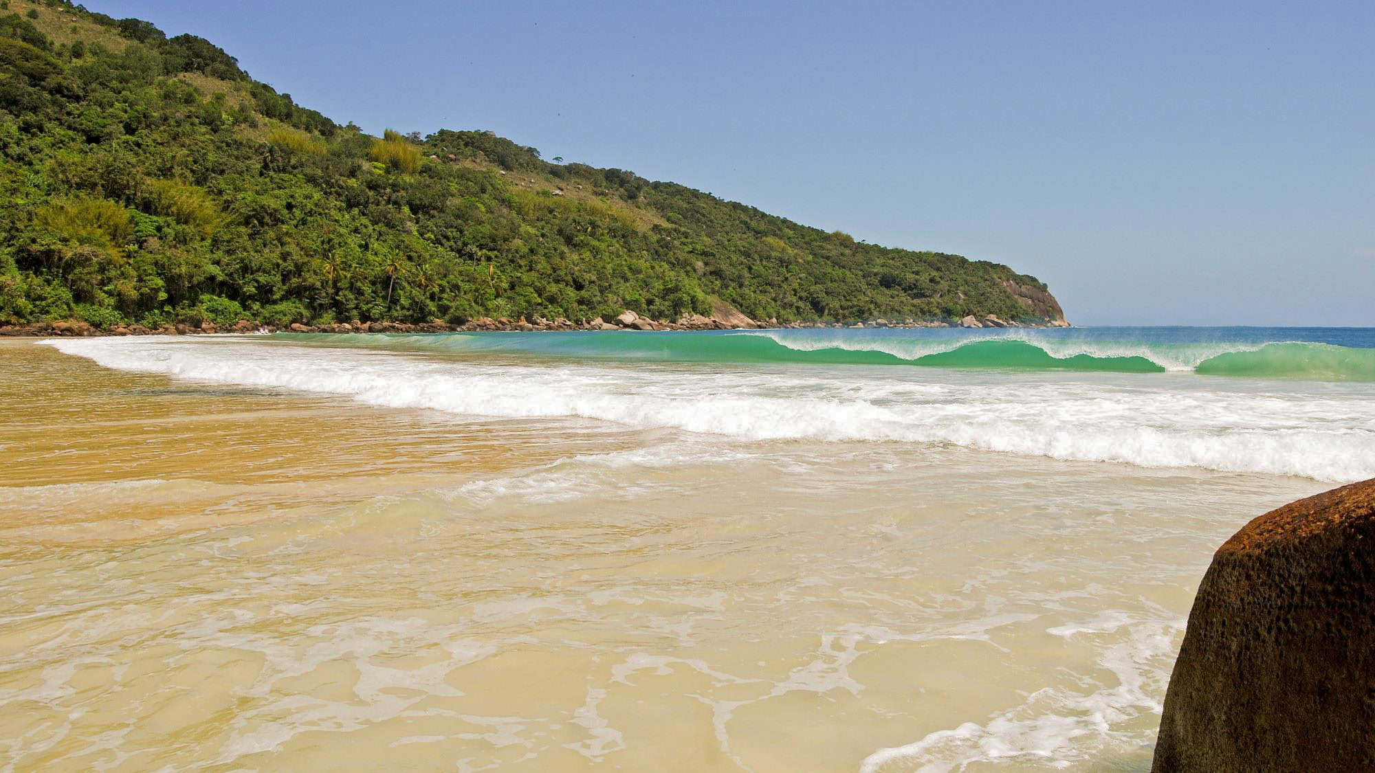 A peaceful tropical beach on Ilha Grande, Brazil, with gentle waves rolling onto the golden shore backed by lush green hills and palm trees.