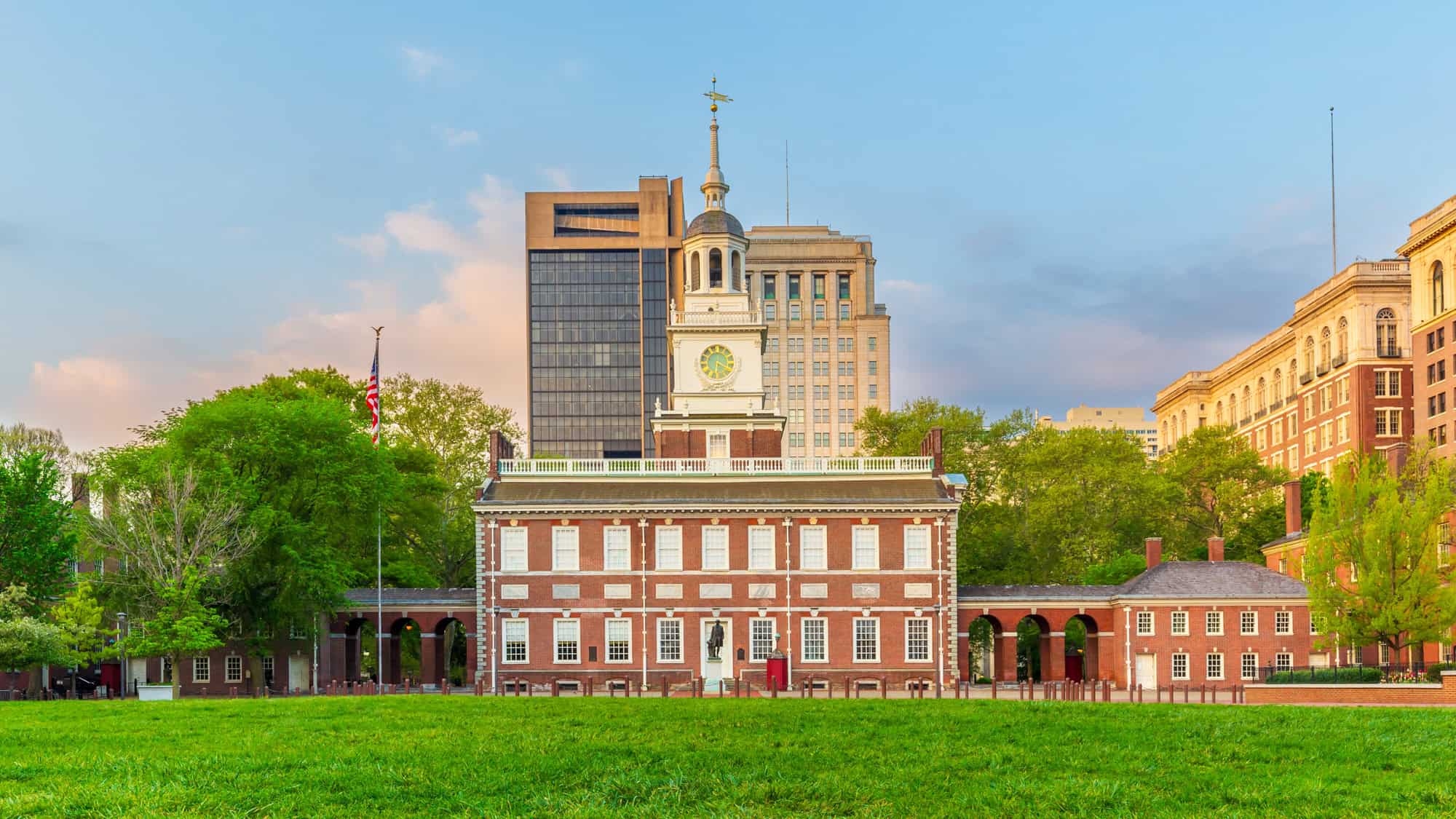 Historic red-brick building with a tall white steeple and clock, set against a backdrop of trees and modern skyscrapers, viewed from a grassy field at sunset.