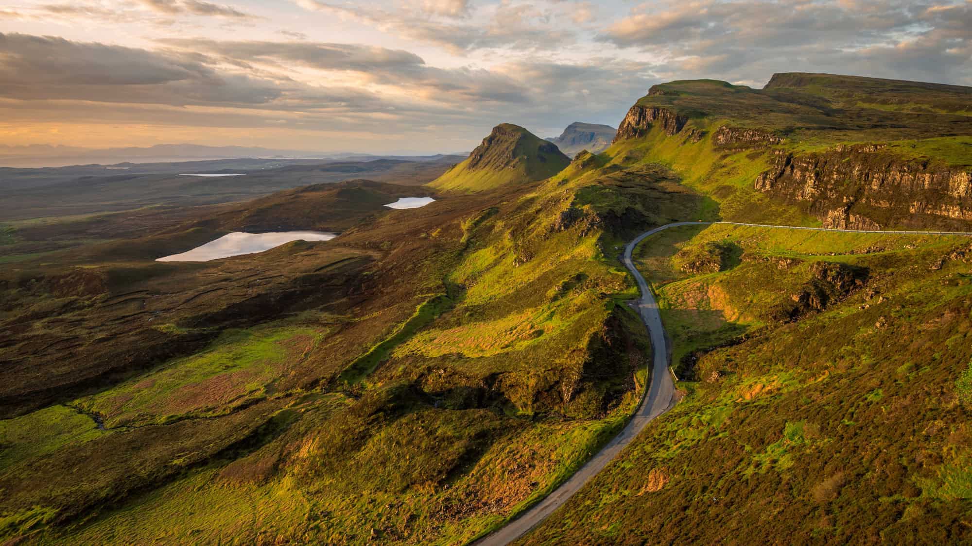 A winding road snakes through the green, rugged hills of the Isle of Skye in Scotland, with dramatic cliffs, rolling moorlands, and shimmering lochs bathed in golden evening light.