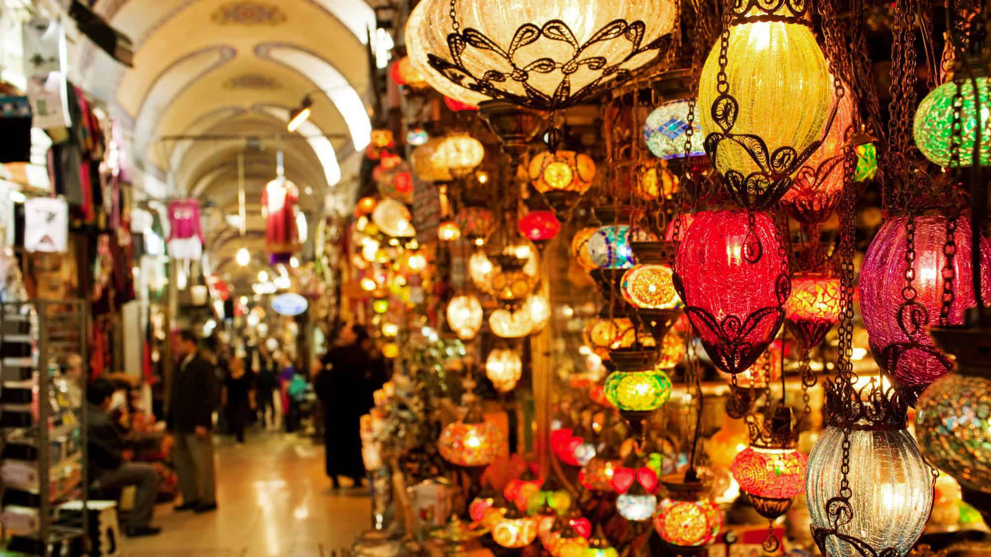A warm, colorful array of hanging mosaic lamps fills the scene at Istanbul’s Grand Bazaar, with arched ceilings above a bustling crowd of shoppers and merchants.